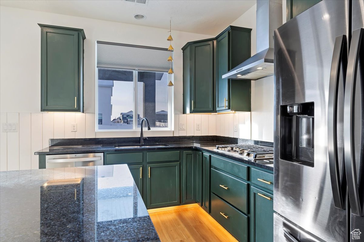 Kitchen with wall chimney exhaust hood, stainless steel appliances, sink, dark stone countertops, and green cabinets