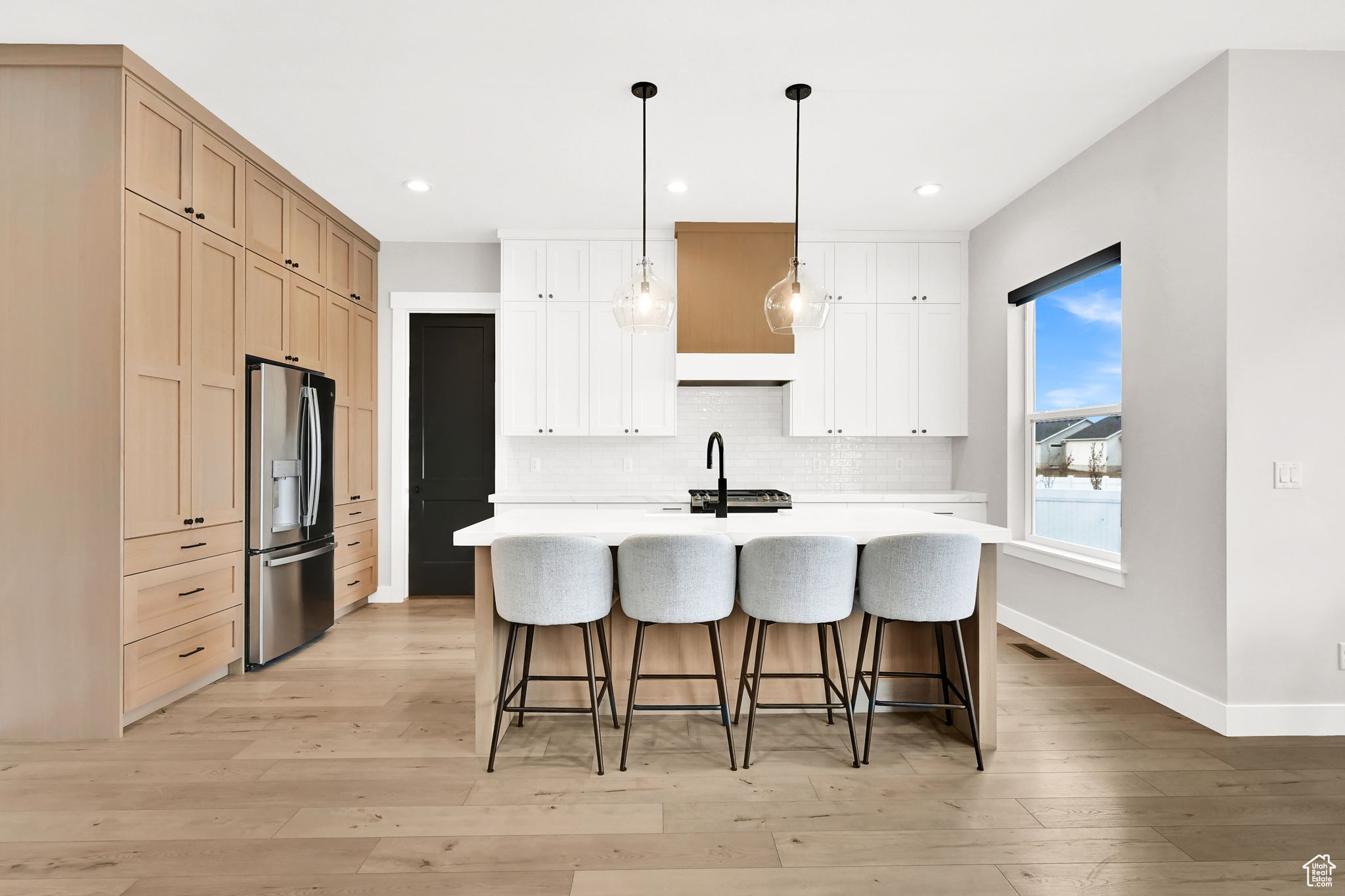 Kitchen with stainless steel fridge, an island with sink, decorative light fixtures, decorative backsplash, and light wood-type flooring