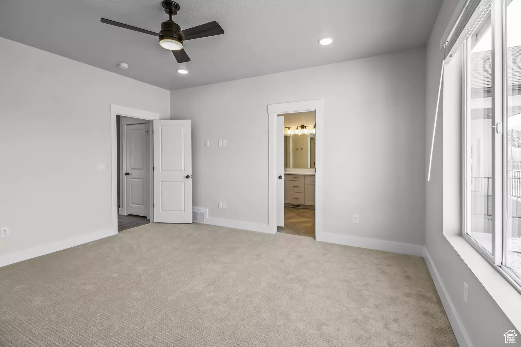 Unfurnished bedroom with multiple windows, light colored carpet, ensuite bath, and ceiling fan