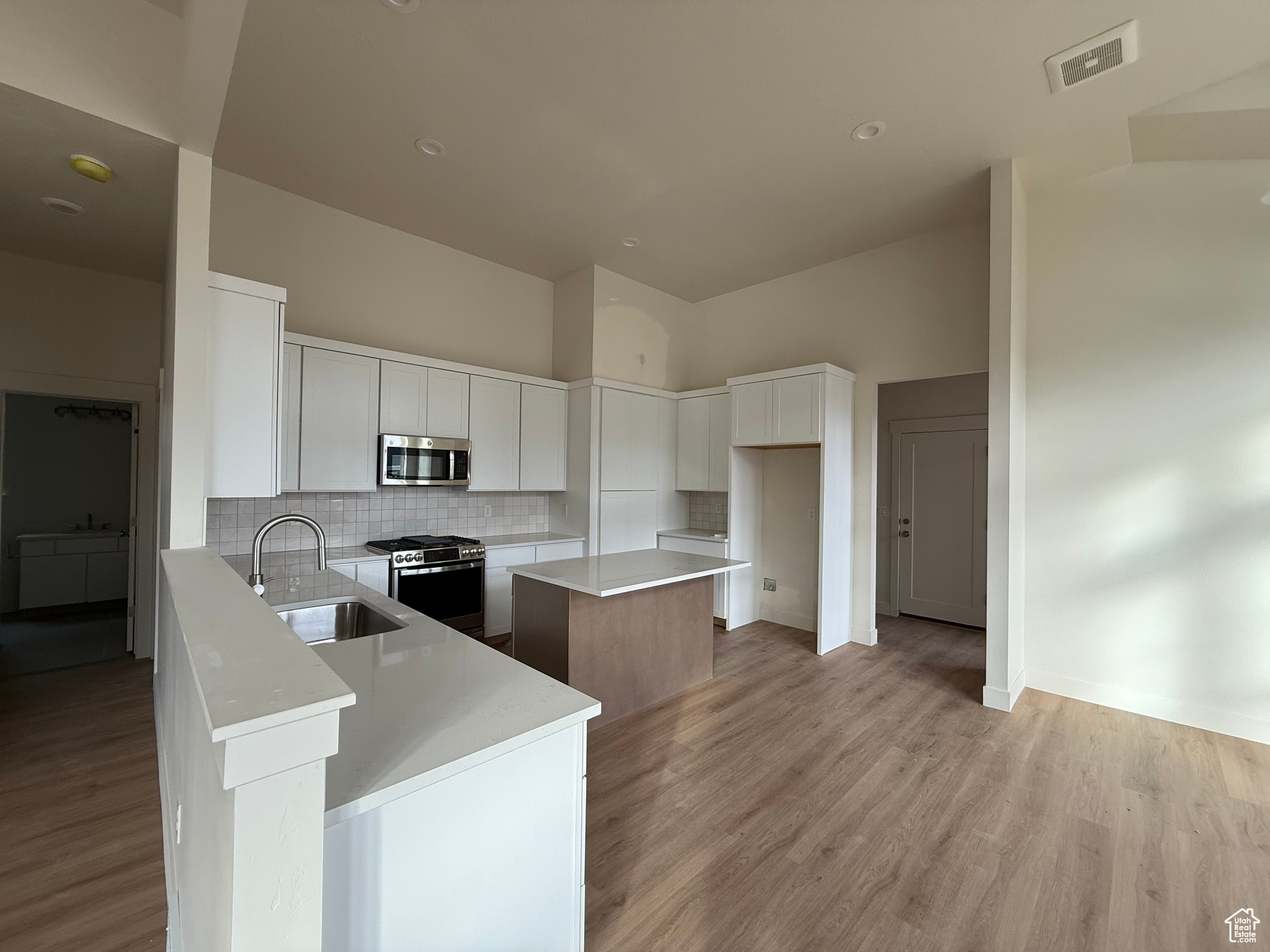 Kitchen with a center island, a high ceiling, white cabinets, sink, and appliances with stainless steel finishes. A pantry is concealed behind the cabinetry to the right of the stove.