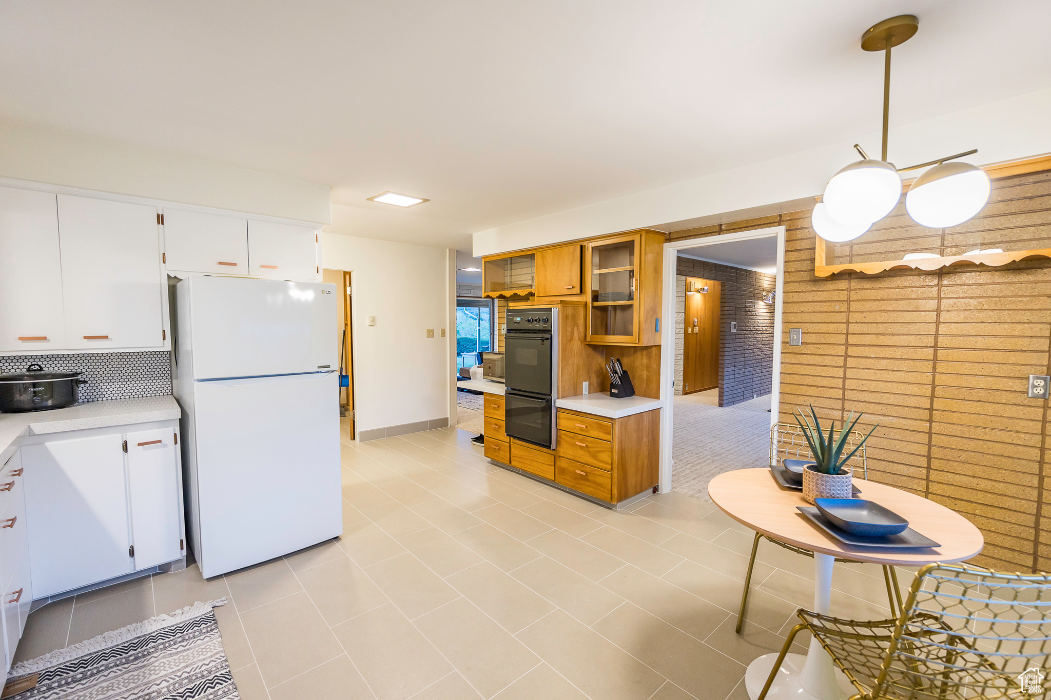 Kitchen with white cabinets, hanging light fixtures, light tile patterned floors, white fridge, and brick wall