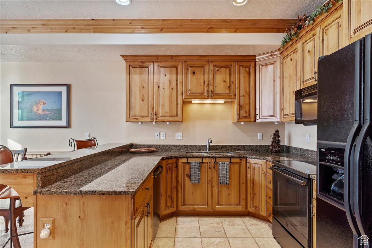 Kitchen featuring sink, kitchen peninsula, dark stone counters, light tile patterned floors, and black appliances