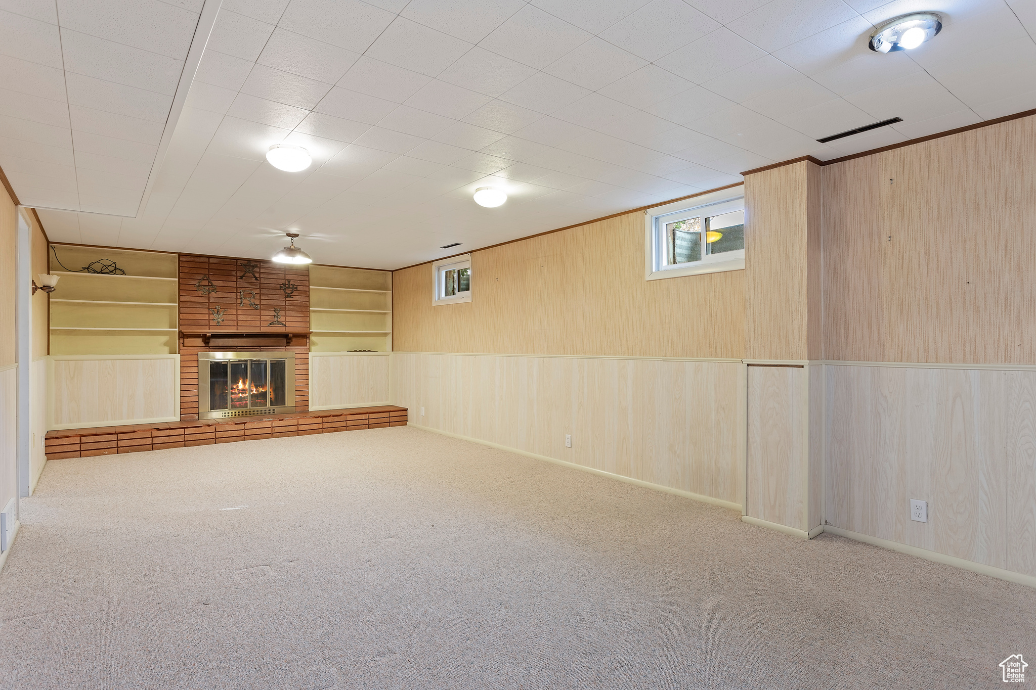 Basement with built in shelves, light carpet, wooden walls, and a brick fireplace