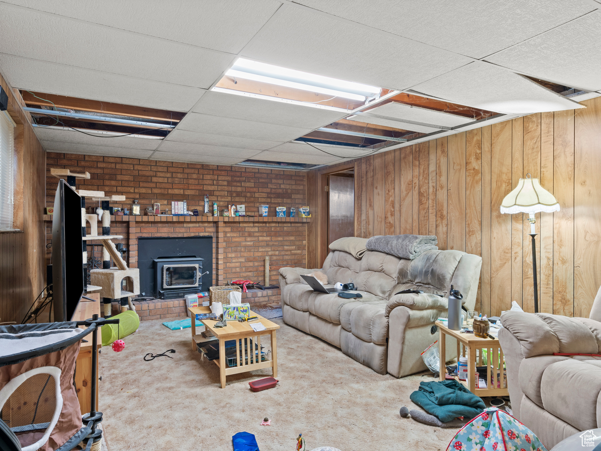 Carpeted living room featuring a paneled ceiling, a wood stove, and wooden walls