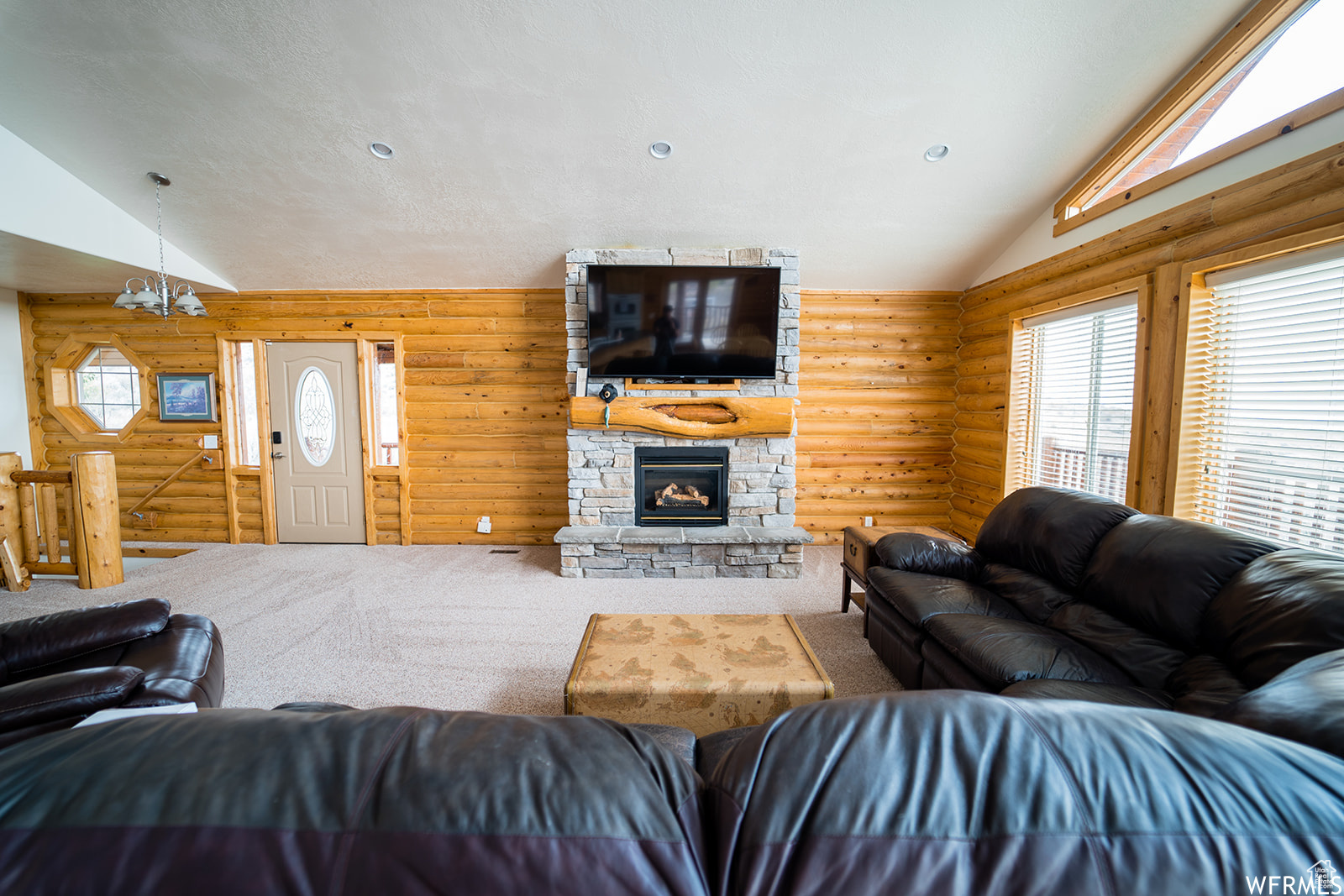 Living room with carpet, lofted ceiling, a stone fireplace, log walls, and a notable chandelier