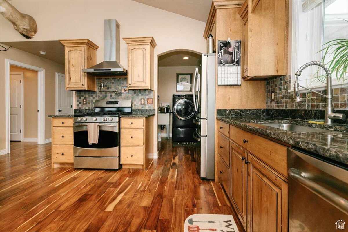 Kitchen with sink, vaulted ceiling, wall chimney exhaust hood, dark stone countertops, and stainless steel appliances