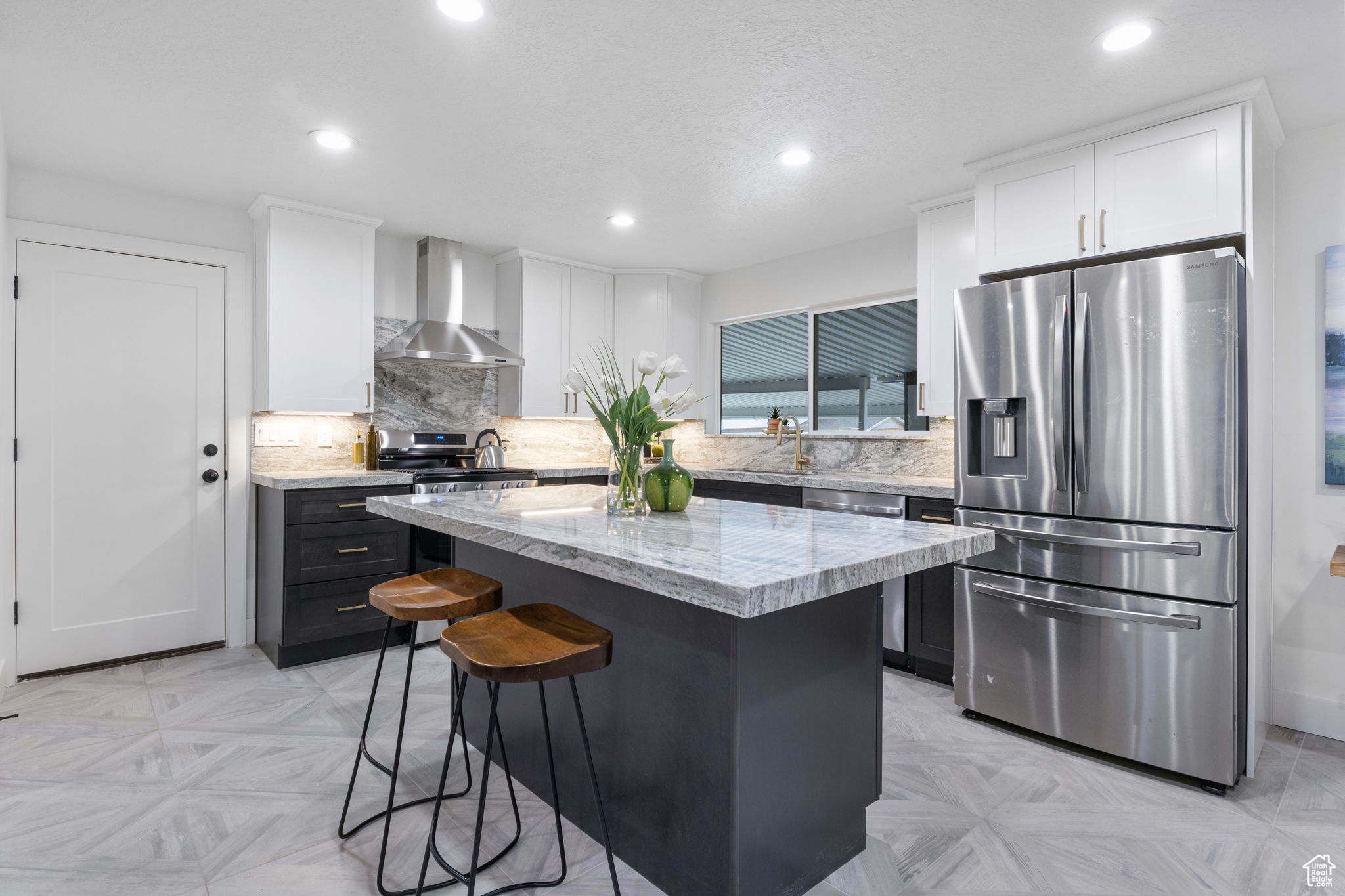 Kitchen with light quartz stone countertops, appliances with stainless steel finishes, wall chimney exhaust hood, a kitchen island, and white and navy blue custom cabinetry