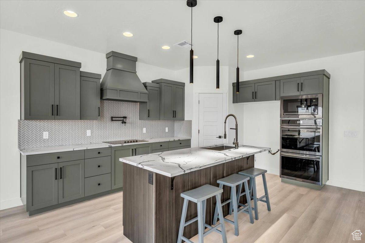 Kitchen with built in microwave, sink, light stone counters, a center island with sink, and custom range hood