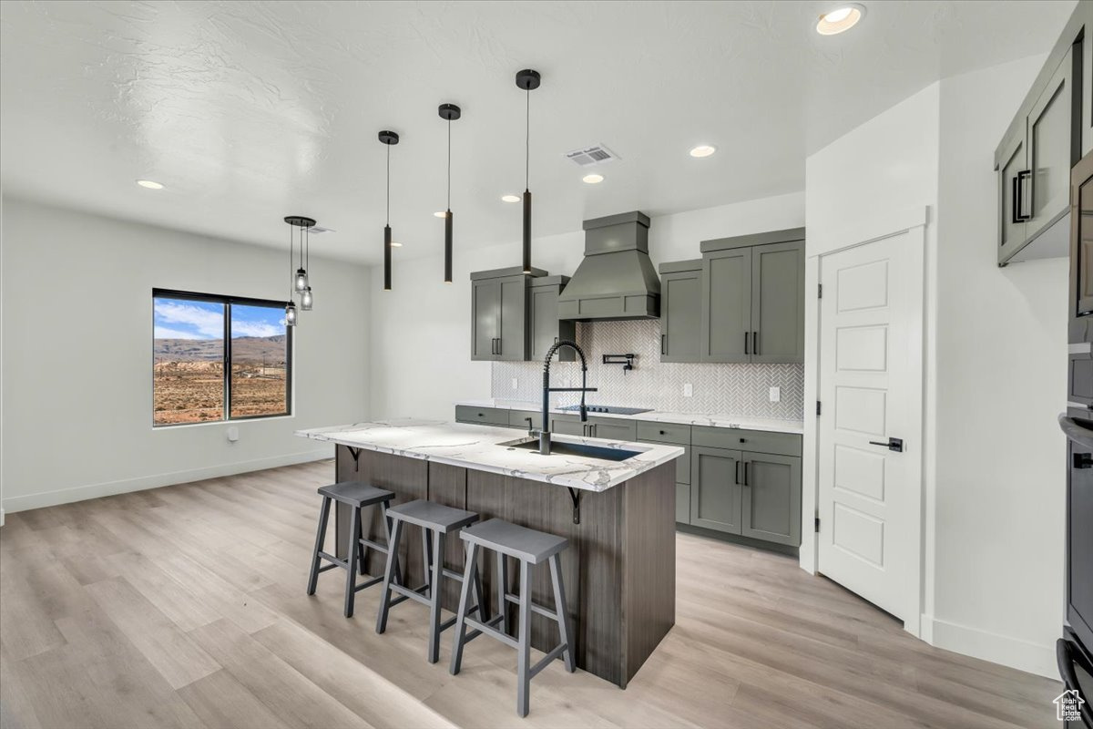 Kitchen featuring a kitchen island with sink, hanging light fixtures, light hardwood / wood-style floors, decorative backsplash, and custom exhaust hood