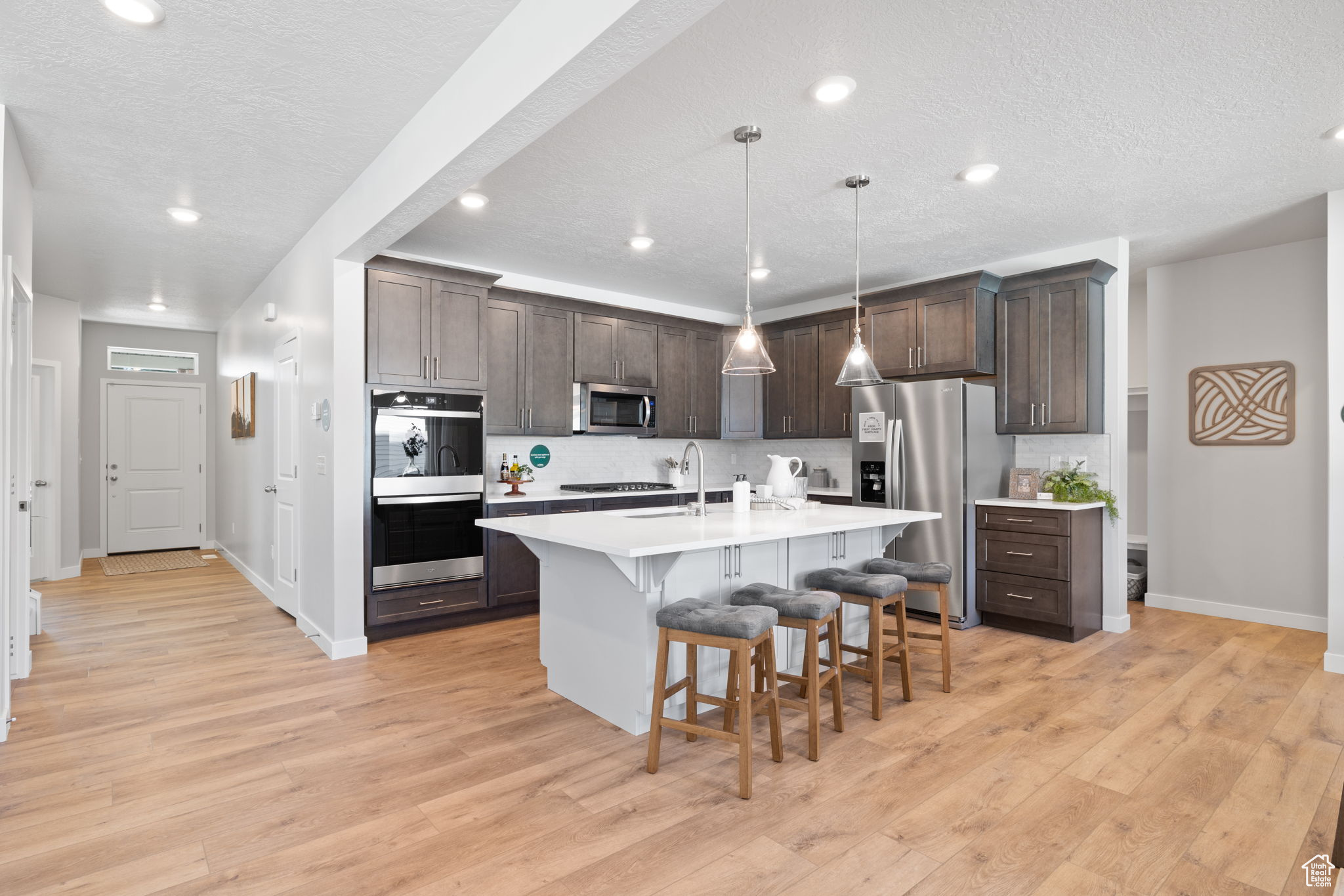 Kitchen featuring dark brown cabinetry, stainless steel appliances, an island with sink, decorative light fixtures, and a kitchen bar