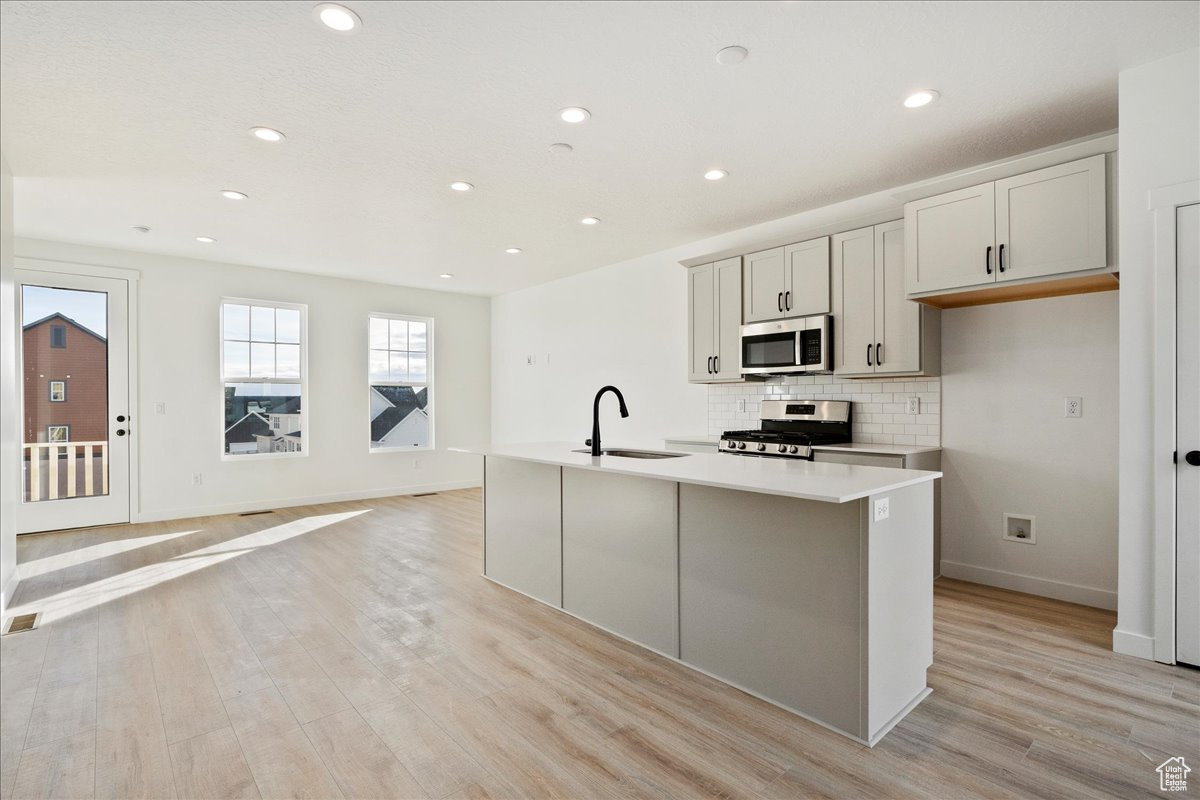 Kitchen with sink, tasteful backsplash, light hardwood / wood-style floors, a center island with sink, and appliances with stainless steel finishes