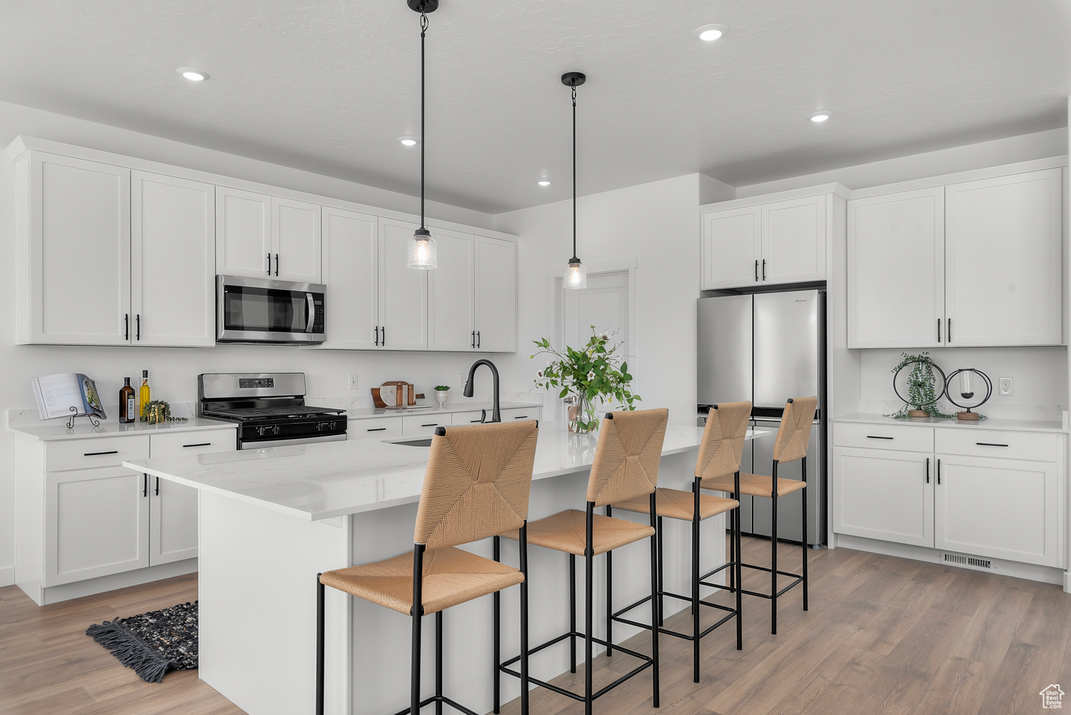 Kitchen with appliances with stainless steel finishes, light wood-type flooring, a kitchen island with sink, white cabinets, and hanging light fixtures