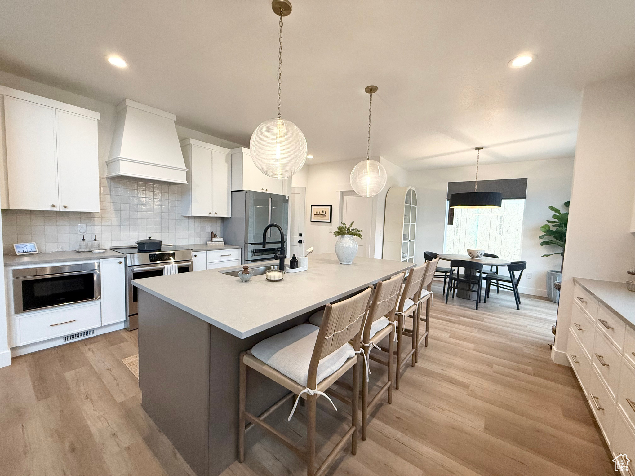 Kitchen with premium range hood, stainless steel appliances, white cabinets, hanging light fixtures, and an island with sink