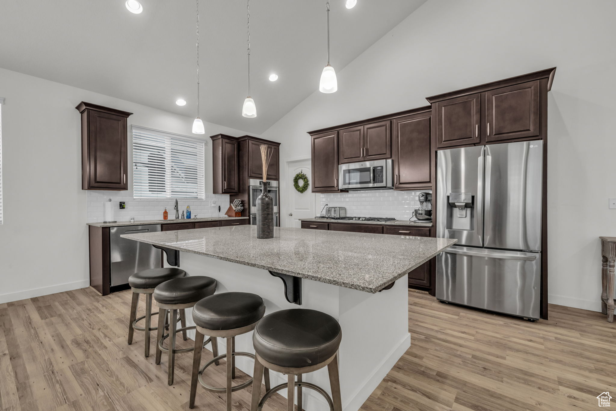 Kitchen with light stone countertops, backsplash, stainless steel appliances, decorative light fixtures, and a center island