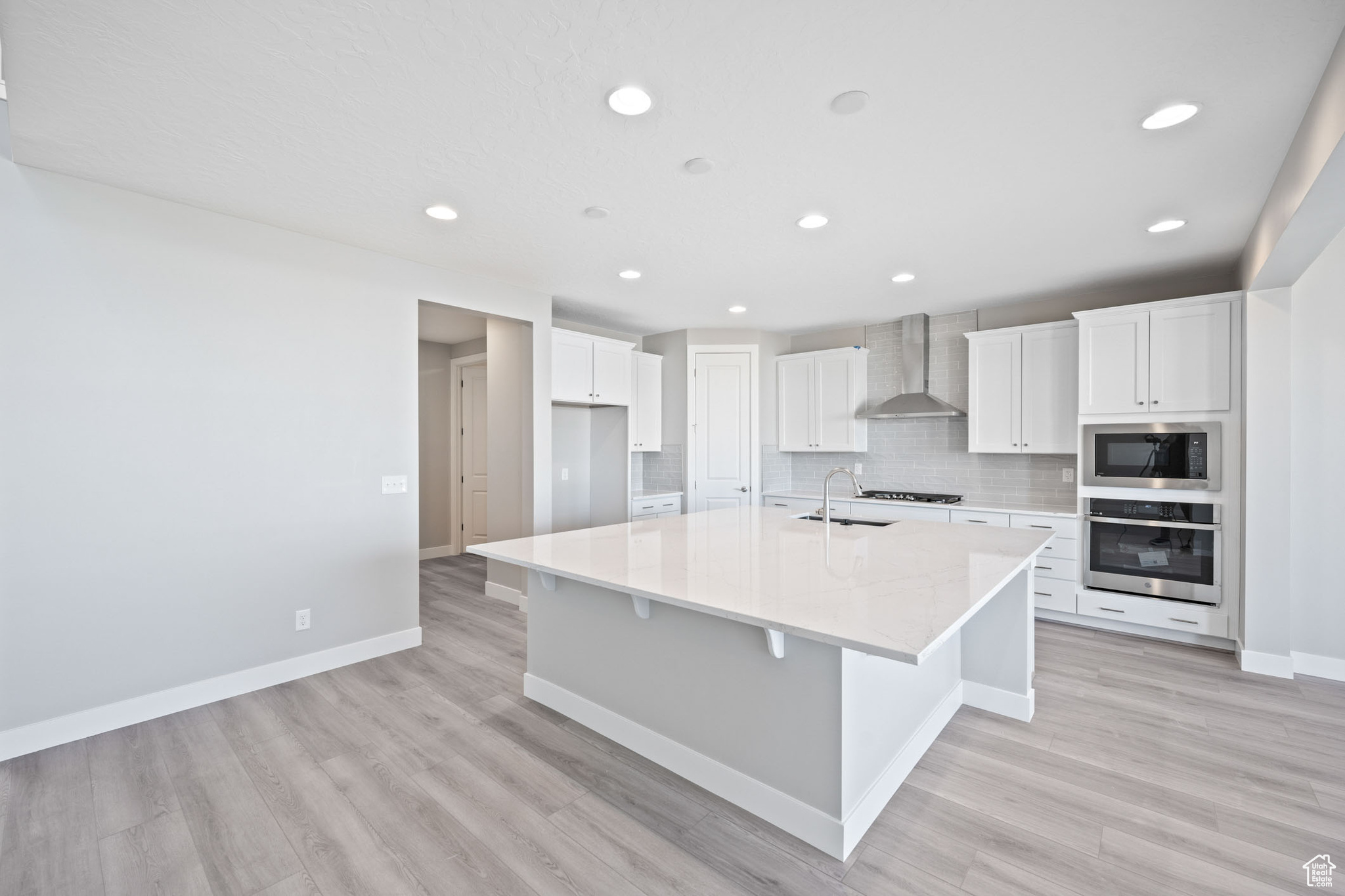 Kitchen featuring white cabinetry, wall chimney exhaust hood, stainless steel appliances, backsplash, and an island with sink