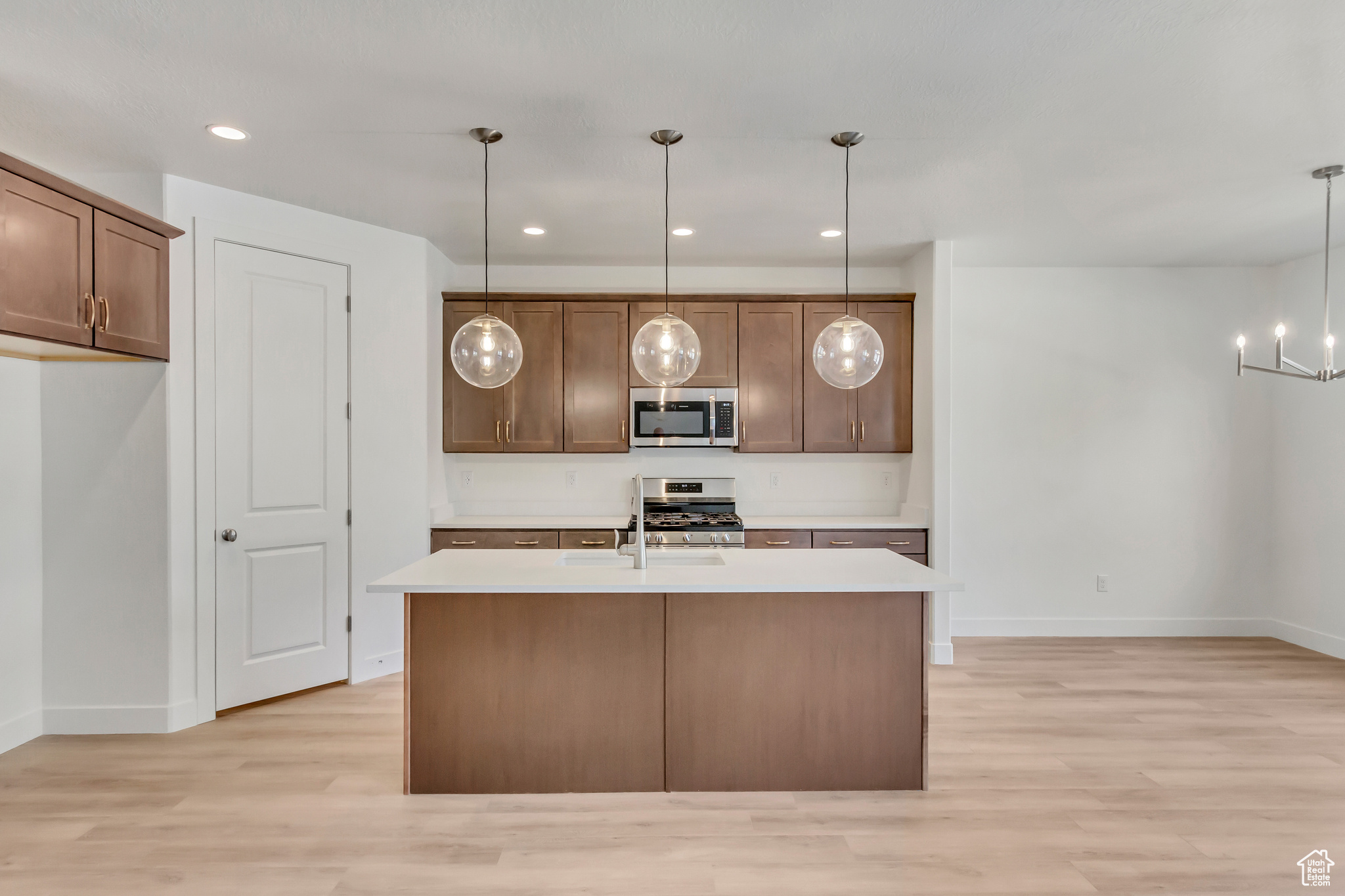 Kitchen with light wood-type flooring, stainless steel appliances, decorative light fixtures, a notable chandelier, and an island with sink