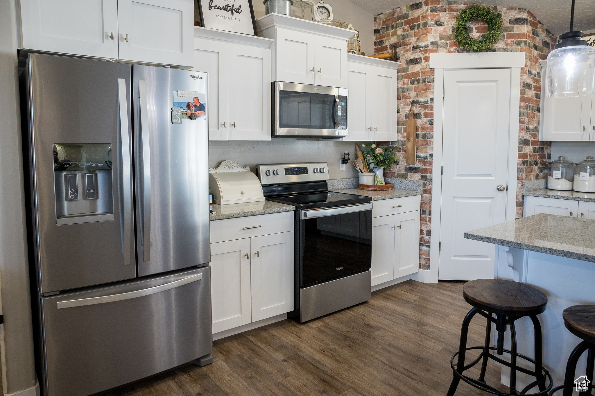 Kitchen with pendant lighting, white cabinets, light stone countertops, stainless steel appliances, and brick wall