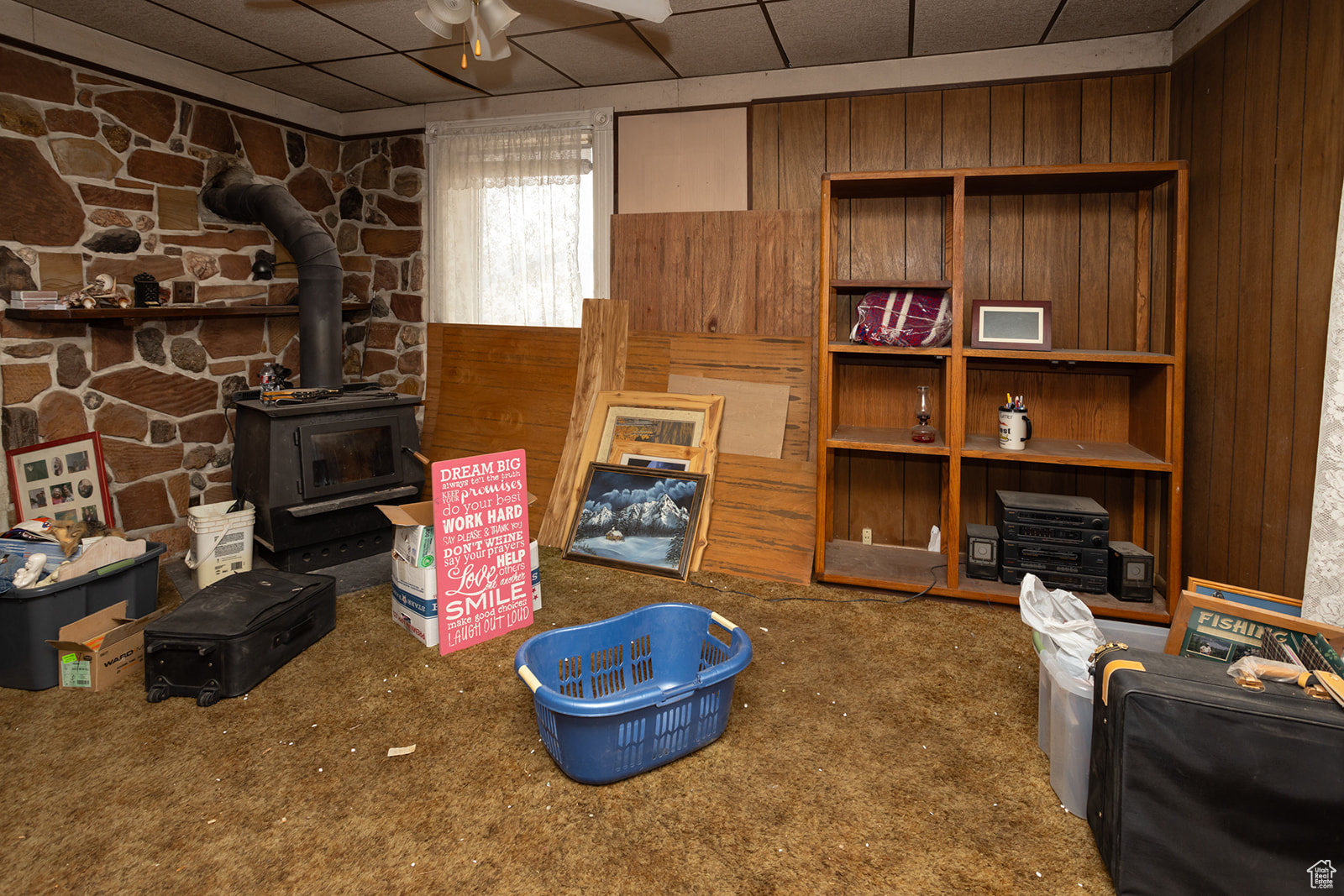 Carpeted living room featuring a wood stove, a paneled ceiling, ceiling fan, and wood walls