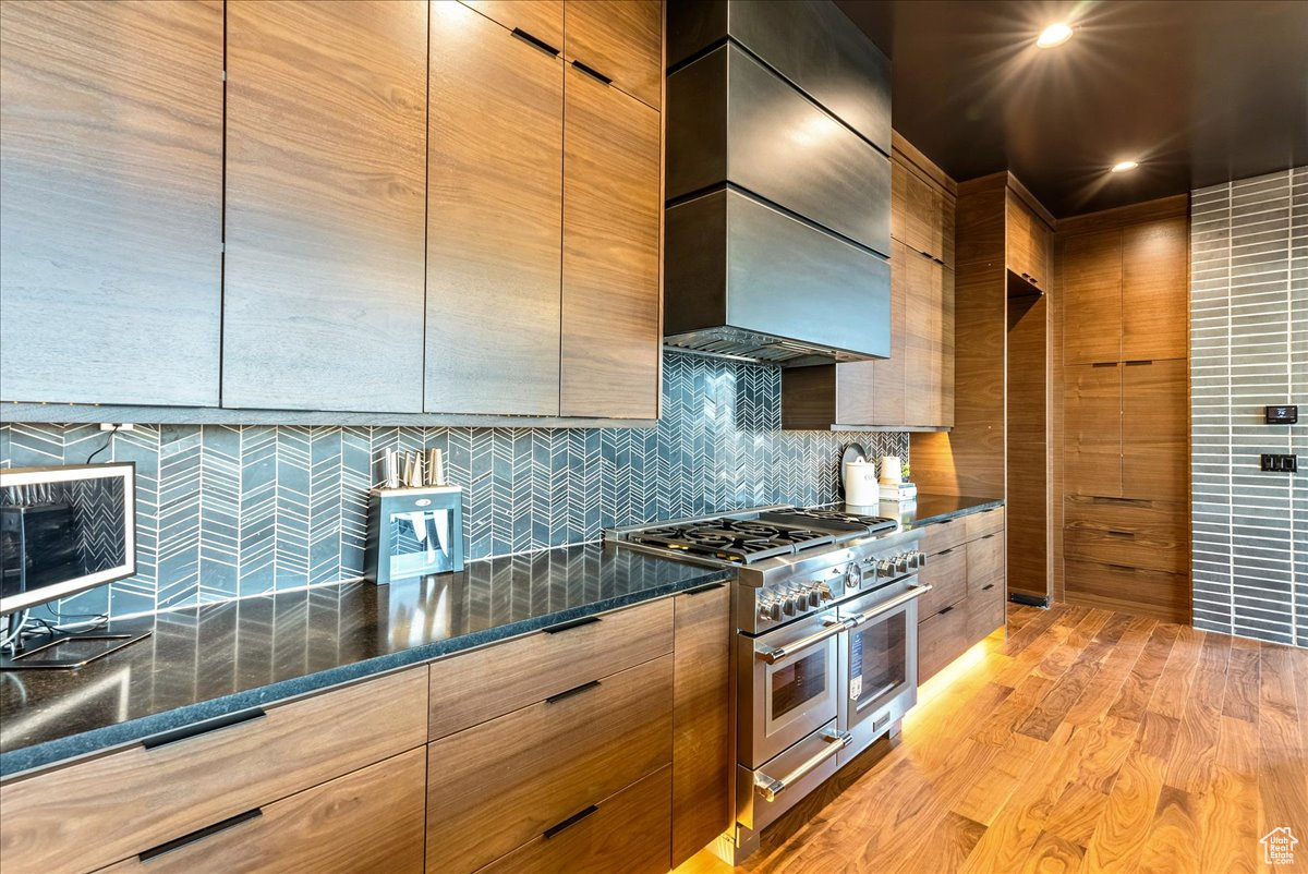 Kitchen featuring decorative backsplash, wall chimney exhaust hood, dark stone counters, light hardwood / wood-style flooring, and double oven range