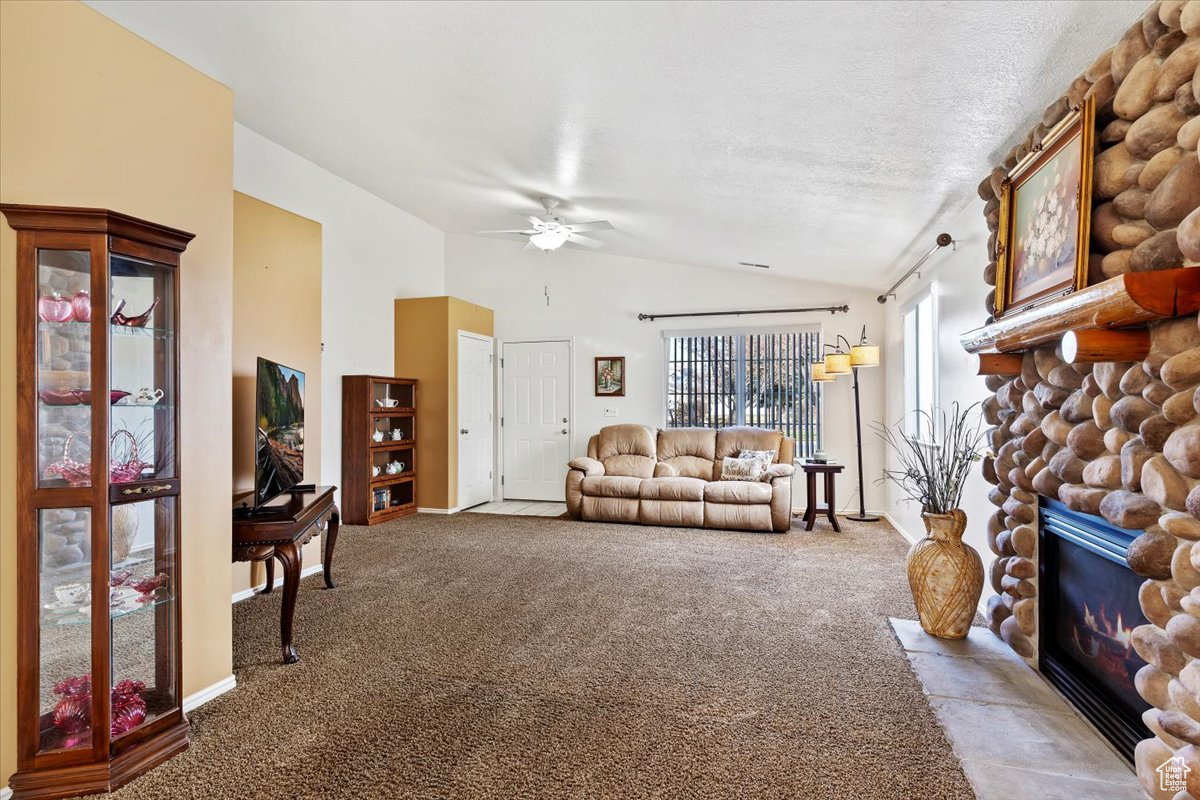 Carpeted living room featuring a textured ceiling, a stone fireplace, ceiling fan, and vaulted ceiling