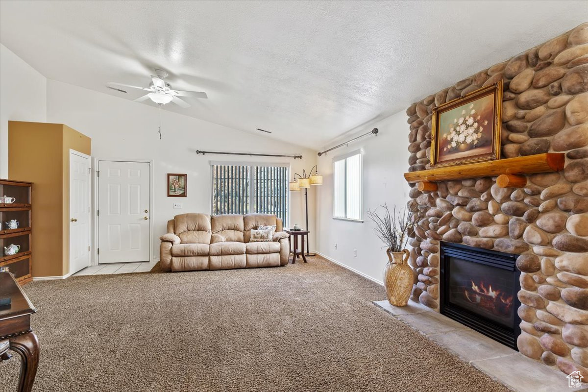 Carpeted living room with ceiling fan, a stone fireplace, lofted ceiling, and a textured ceiling