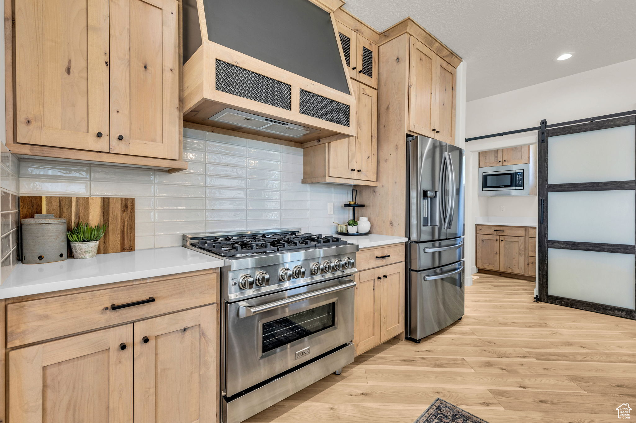 Kitchen featuring light brown cabinets, stainless steel appliances, a barn door, ventilation hood, and backsplash