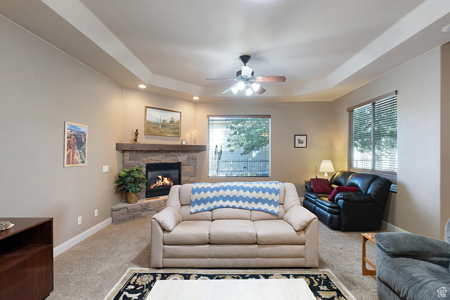 Carpeted living room with ceiling fan, a stone fireplace, and a tray ceiling