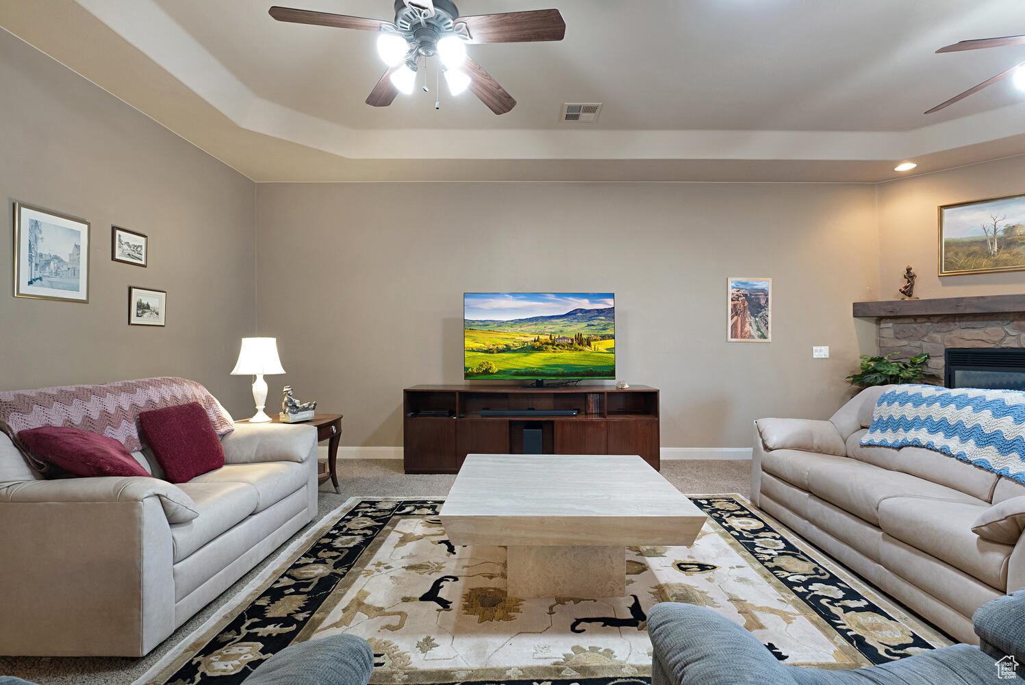 Living room with carpet, ceiling fan, a stone fireplace, and a tray ceiling