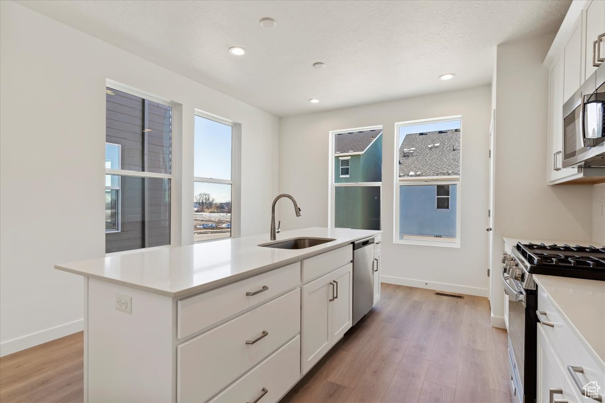 Kitchen with a center island with sink, sink, light wood-type flooring, appliances with stainless steel finishes, and white cabinetry