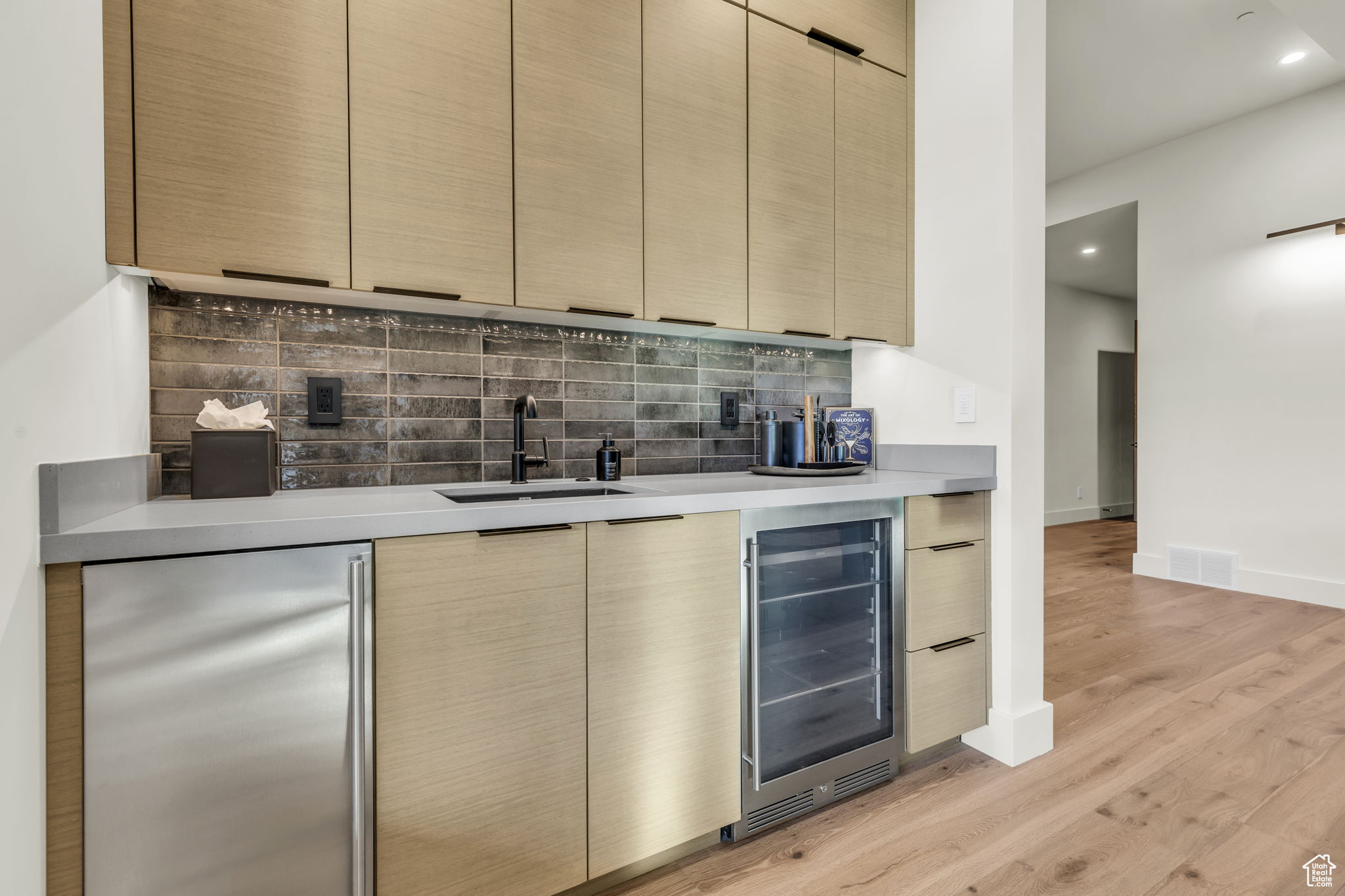 Bar with light wood-type flooring, backsplash, beverage cooler, sink, and fridge