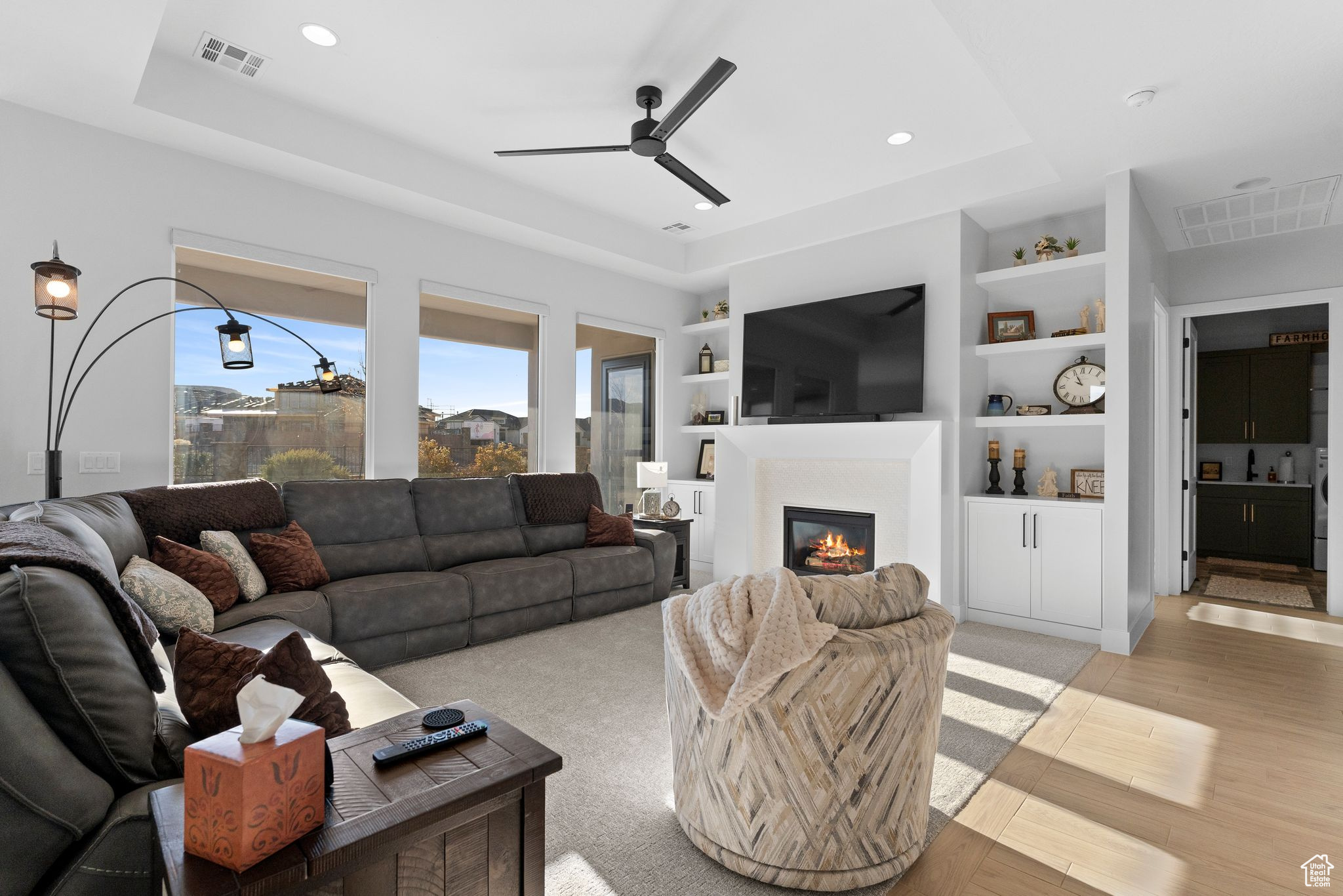 Living room with a raised ceiling, light hardwood / wood-style flooring, built in features, ceiling fan, remote blinds and gas fireplace.