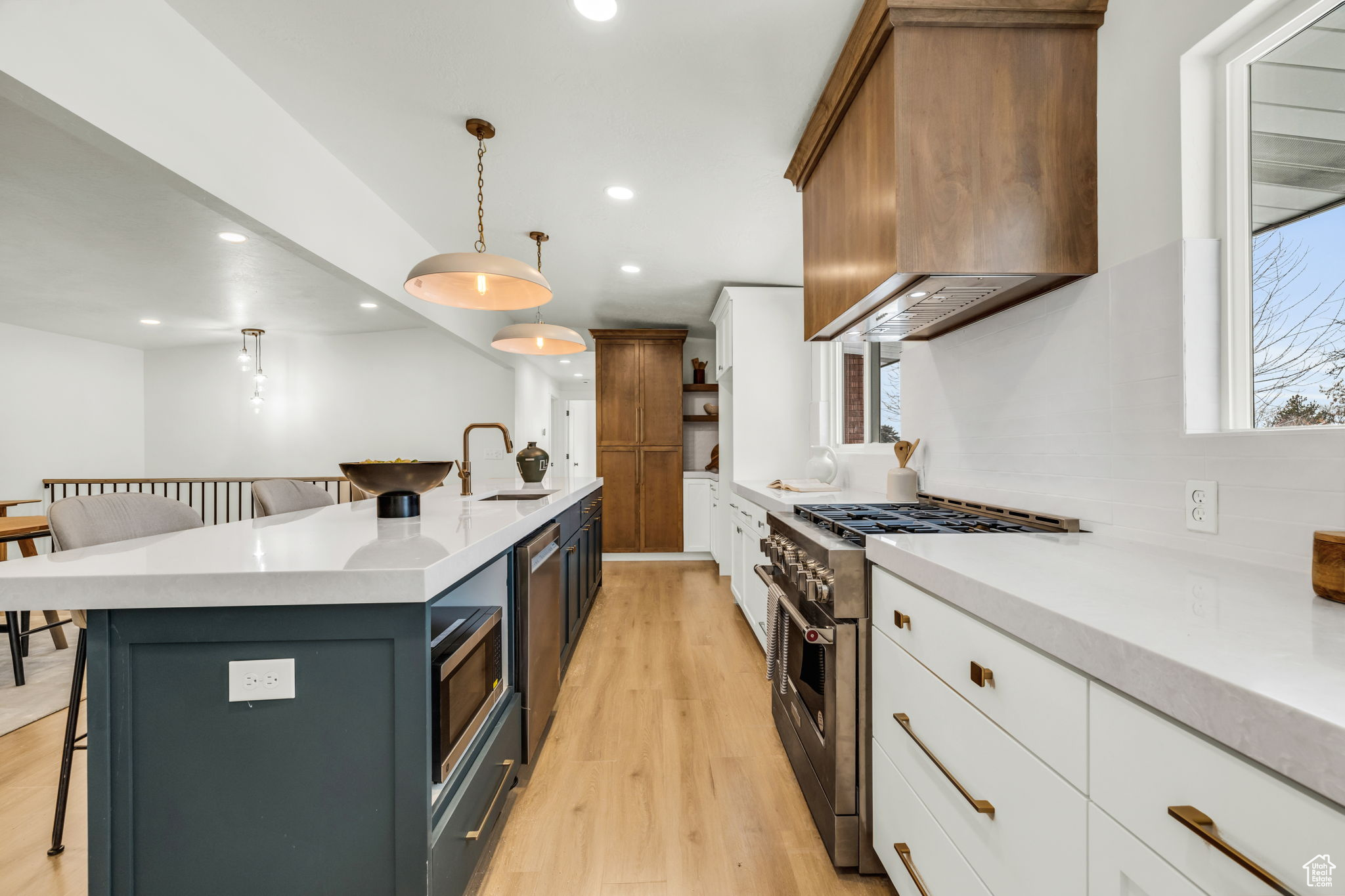 Kitchen featuring pendant lighting, a kitchen island with sink, wall chimney exhaust hood, appliances with stainless steel finishes, and white cabinetry