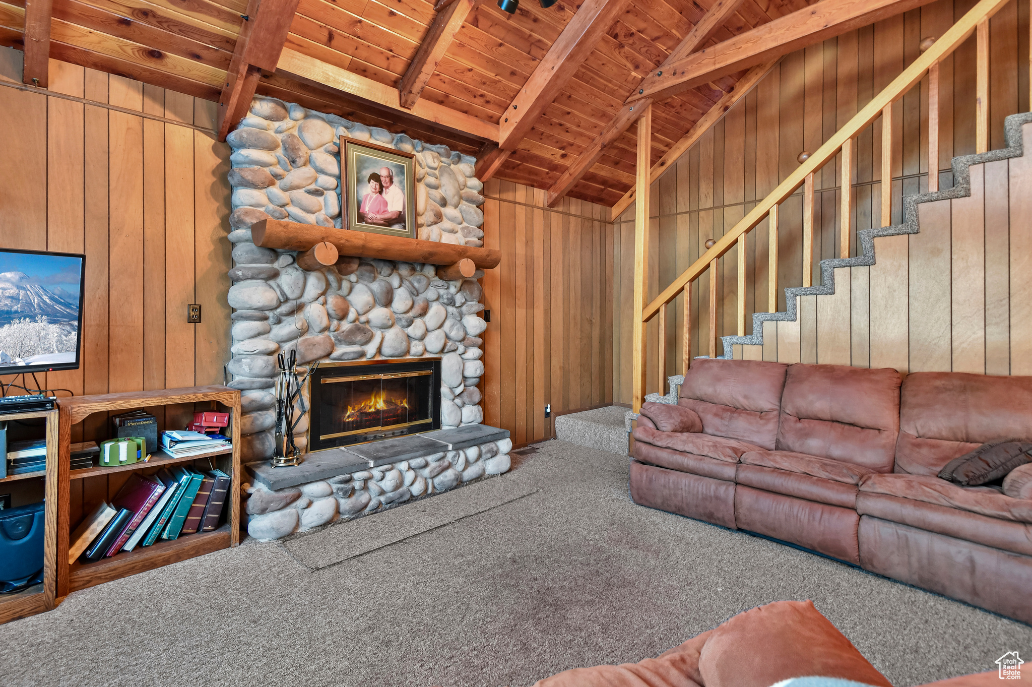 Living room featuring carpet, a stone fireplace, wooden walls, beam ceiling, and wood ceiling