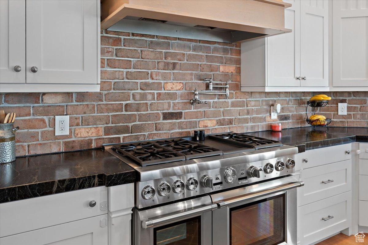 Kitchen featuring decorative backsplash, wall chimney range hood, double oven range, dark stone countertops, and white cabinetry