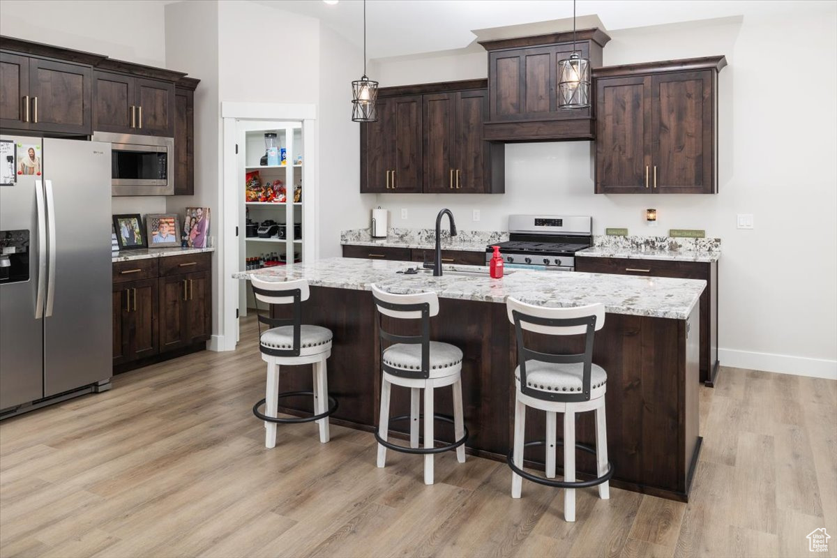 Kitchen featuring stainless steel appliances, an island with sink, hanging light fixtures, light stone counters, and dark brown cabinets