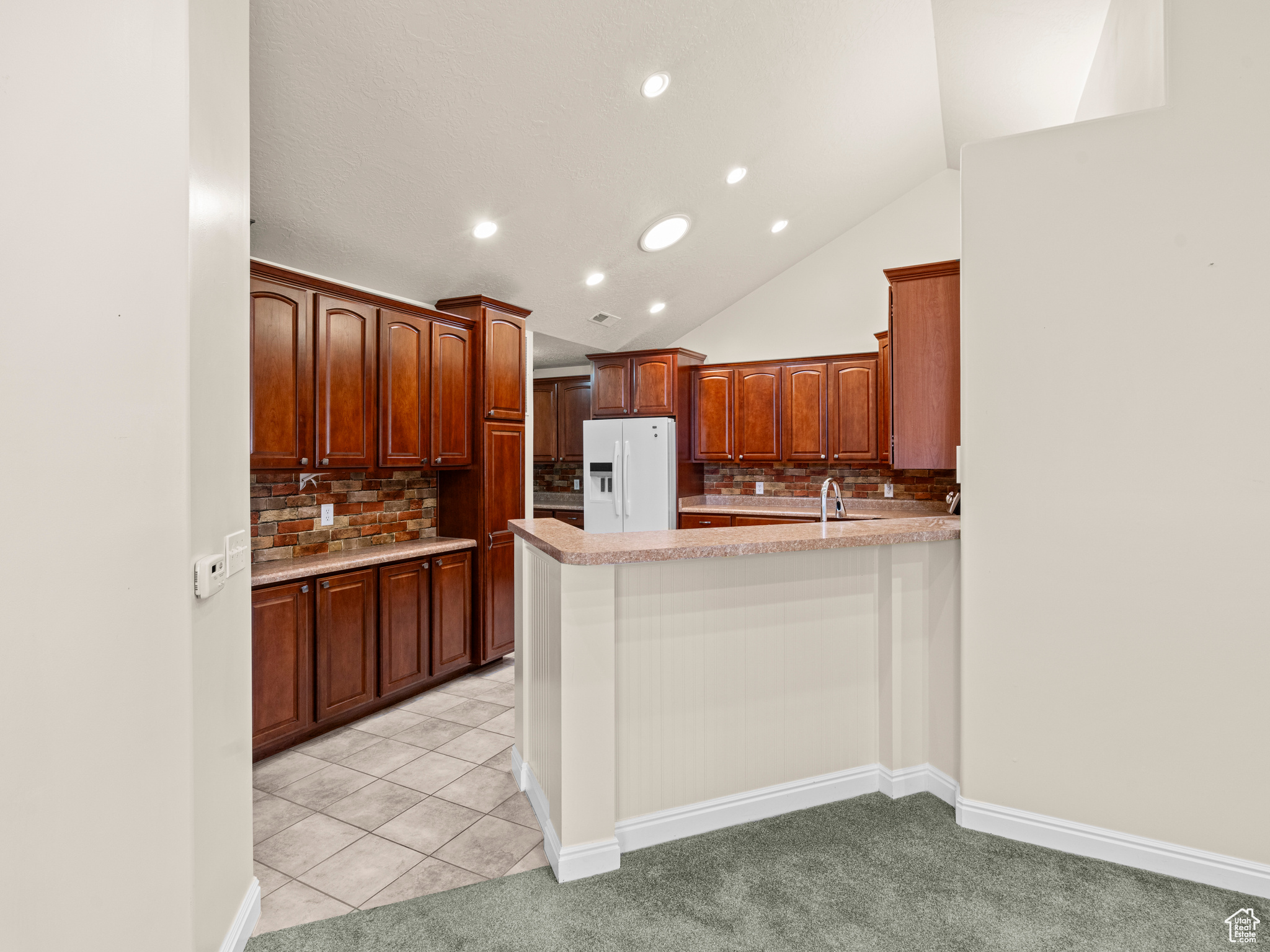 Kitchen featuring vaulted ceiling, white refrigerator with ice dispenser, decorative backsplash, kitchen peninsula, and light tile patterned flooring