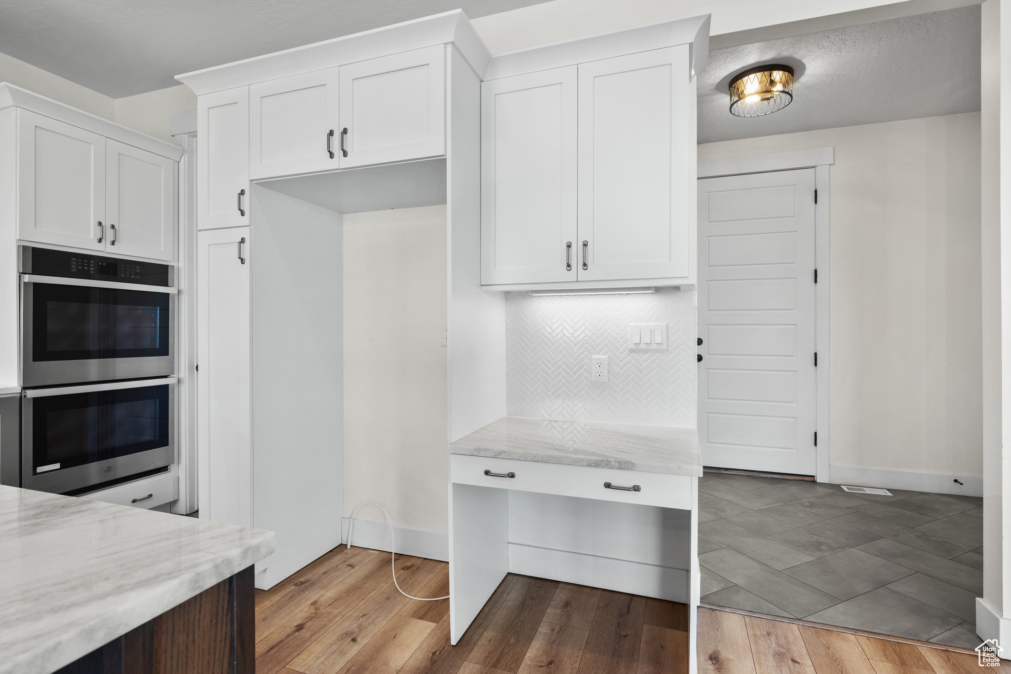 Kitchen with white cabinets, backsplash, light stone countertops, stainless steel double oven, and light hardwood / wood-style flooring