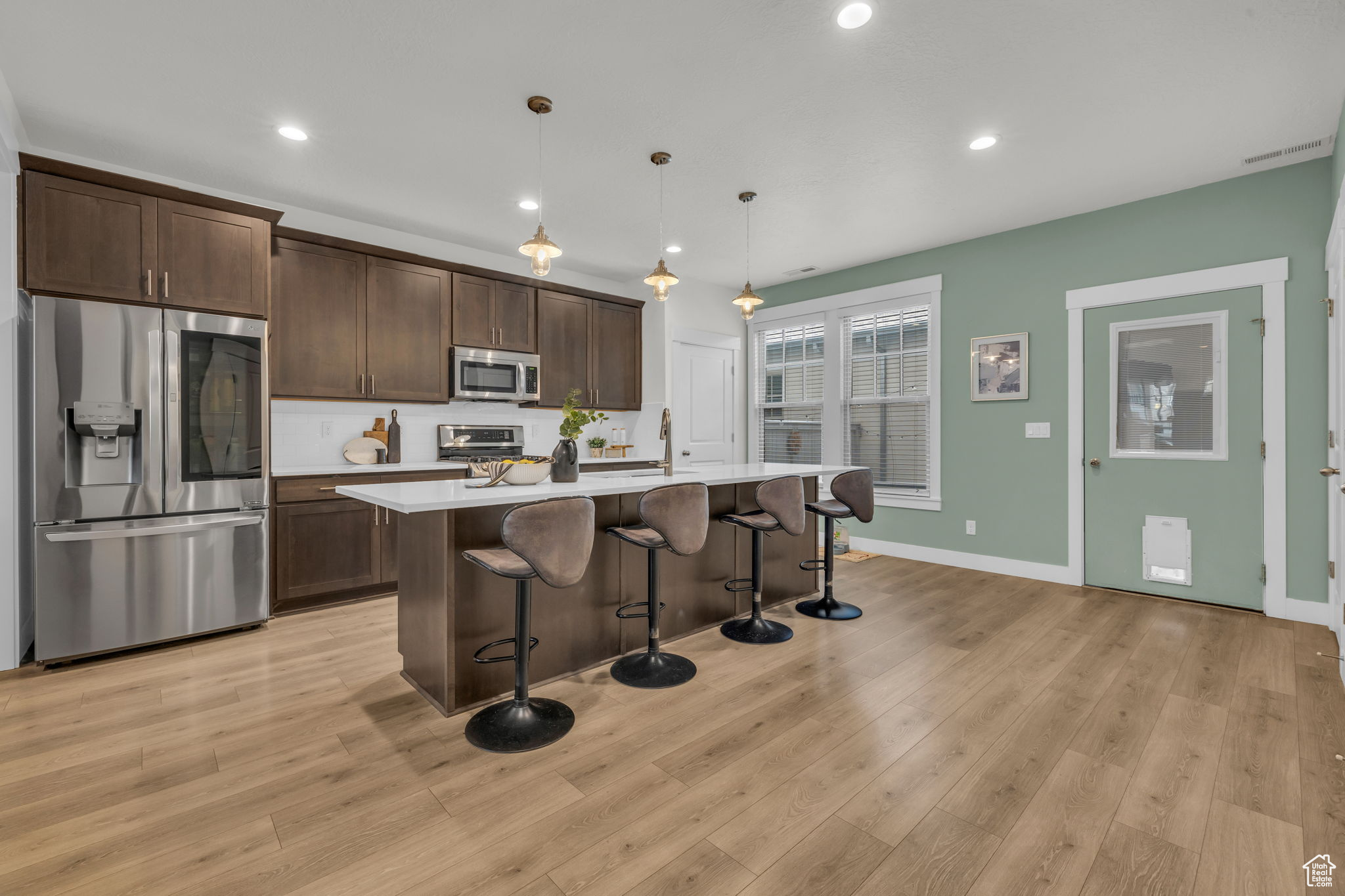 Kitchen with appliances with stainless steel finishes, a center island with sink, light wood-type flooring, and decorative light fixtures