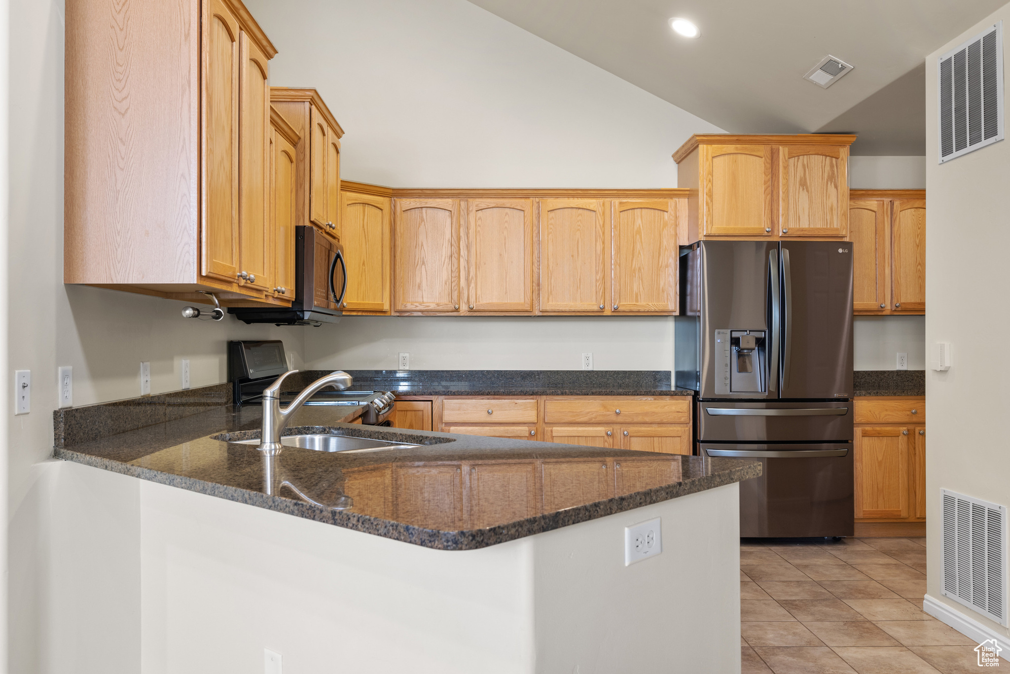 Kitchen with light tile patterned flooring, sink, dark stone countertops, refrigerator with ice dispenser, and kitchen peninsula