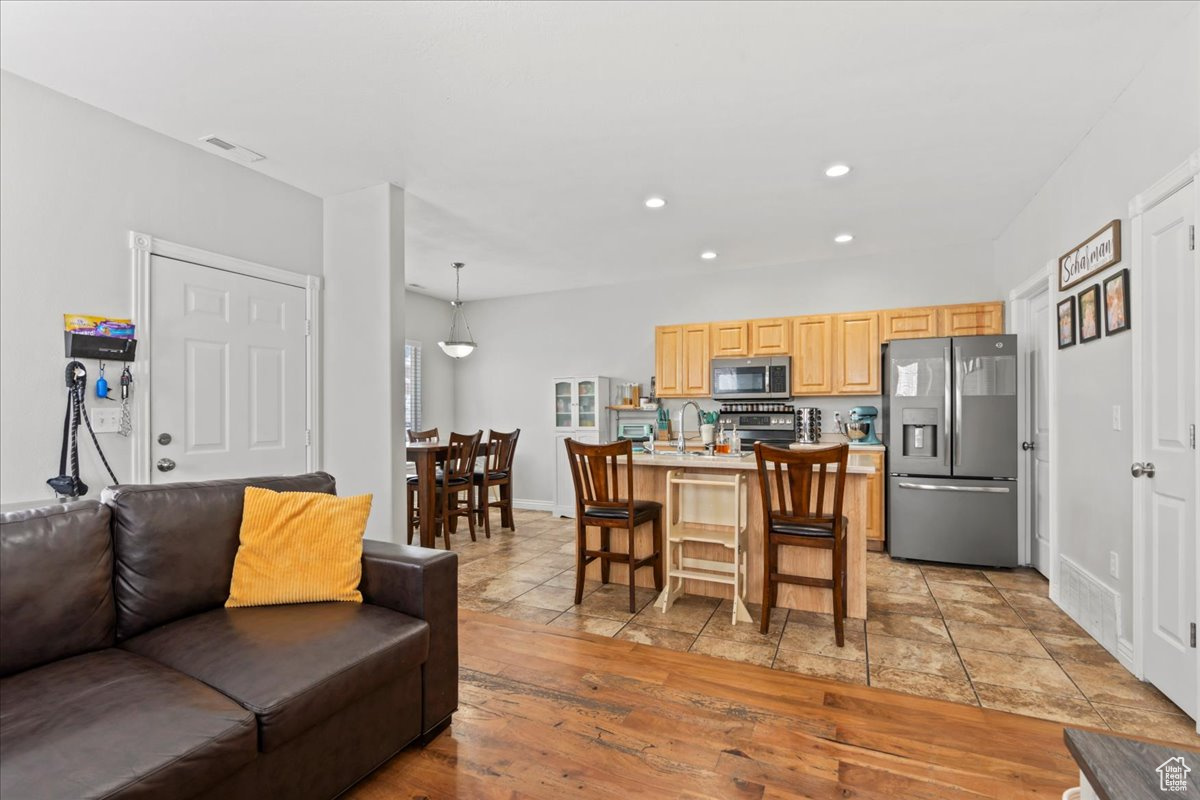 Interior space featuring a breakfast bar area, appliances with stainless steel finishes, hanging light fixtures, a center island with sink, and light brown cabinetry