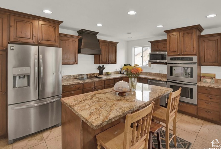 Kitchen with a kitchen island, a breakfast bar, stainless steel appliances, light stone countertops, and custom range hood