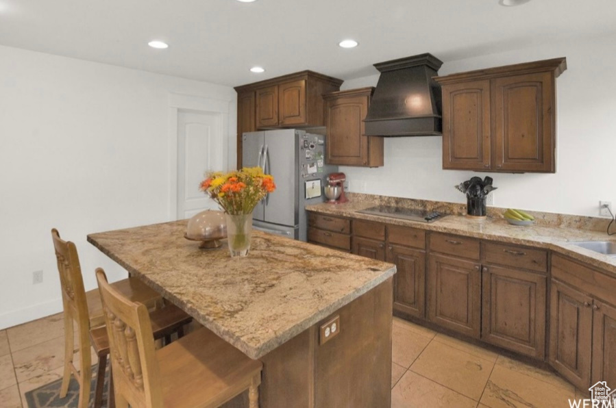 Kitchen with a breakfast bar, stainless steel refrigerator, a center island, custom range hood, and black electric cooktop