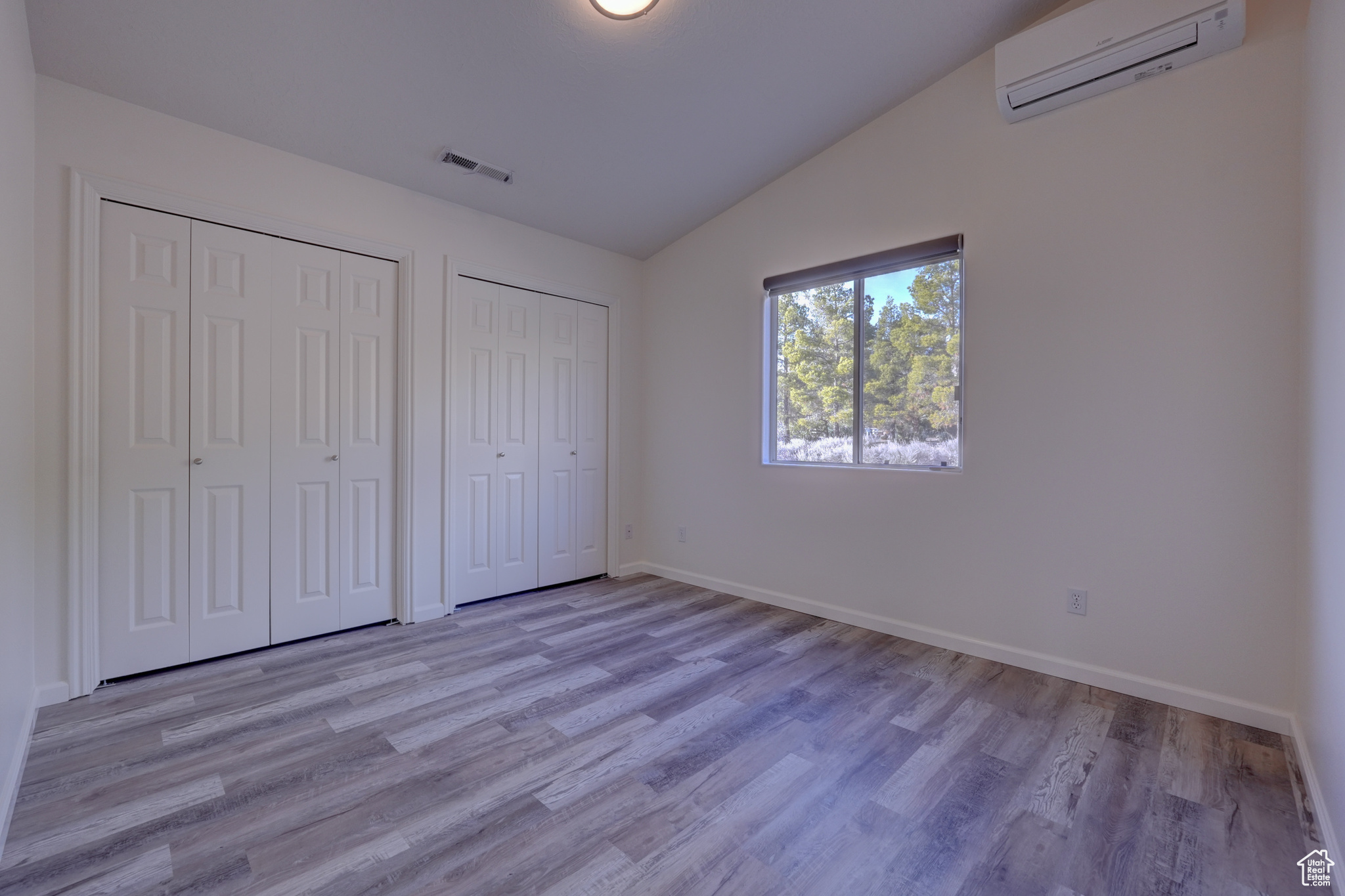 Unfurnished bedroom with lofted ceiling, a wall mounted AC, light wood-type flooring, and two closets