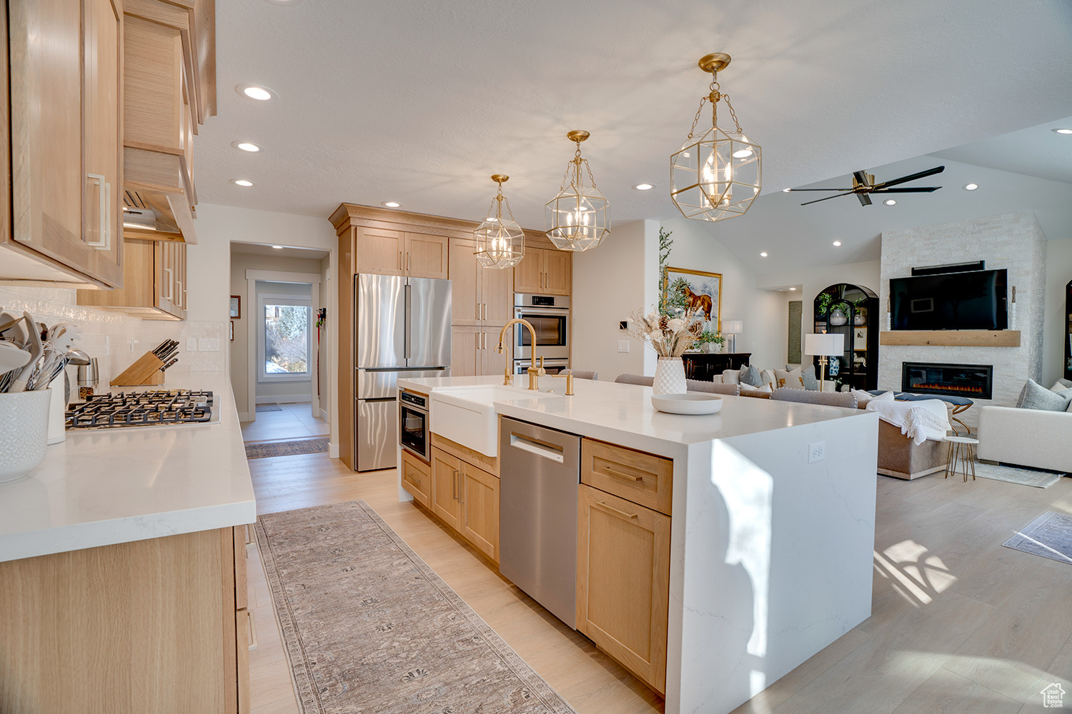 Kitchen with decorative light fixtures, a center island with sink, light brown cabinets, a large fireplace, and stainless steel appliances