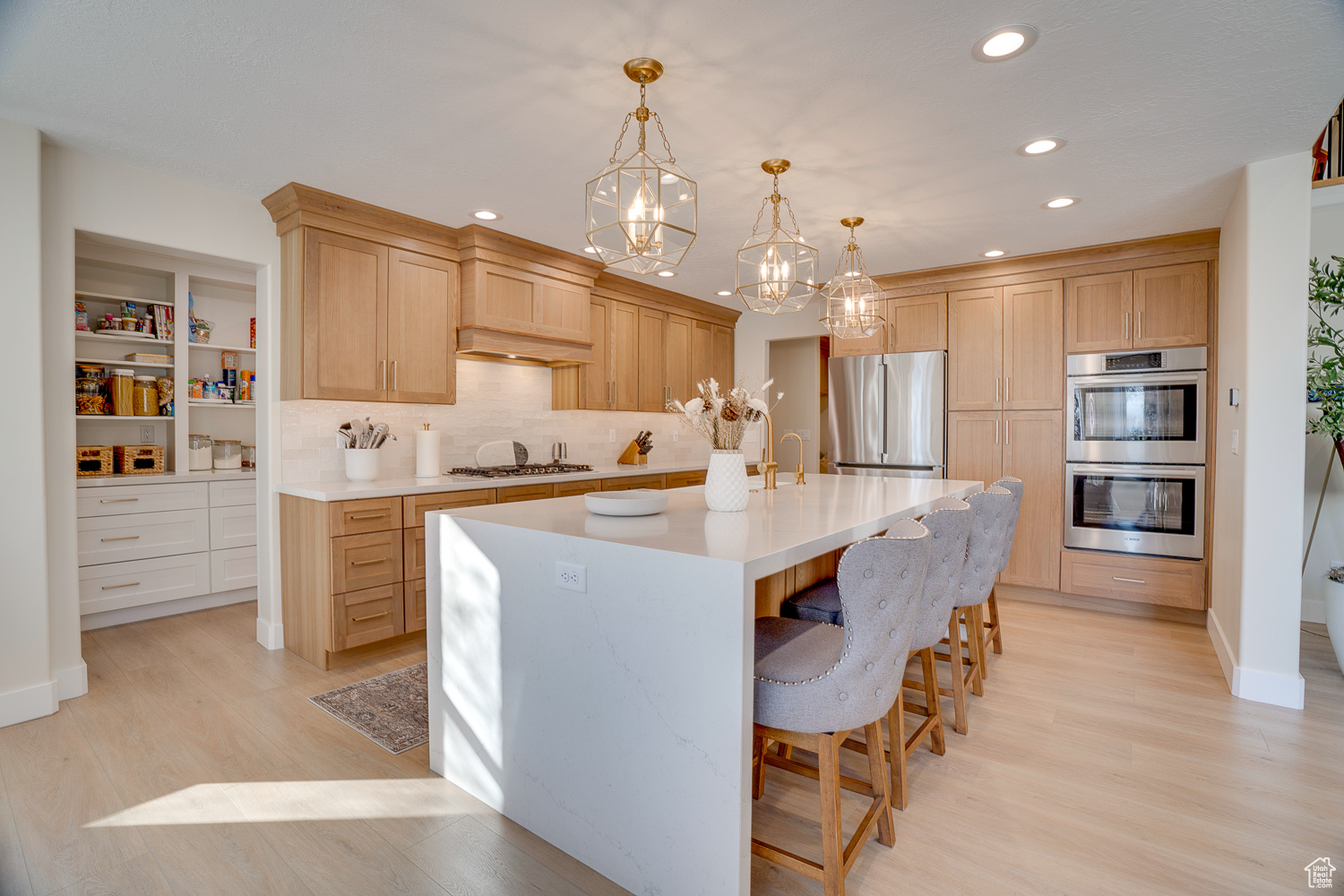 Kitchen with pendant lighting, appliances with stainless steel finishes, light hardwood / wood-style floors, an island with sink, and light brown cabinets