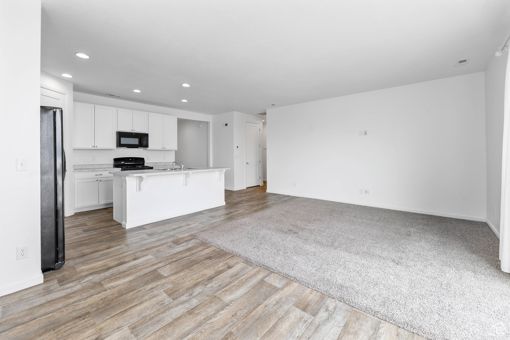 View of kitchen featuring a breakfast bar area, white cabinetry, light wood-type flooring, a kitchen island with sink, and black appliances along with the living room are all from the view point from the dining room