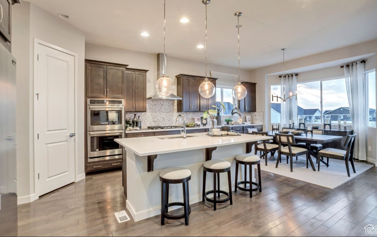 Kitchen with wall oven/microwave, decorative light fixtures, an island with sink, and wall chimney exhaust hood