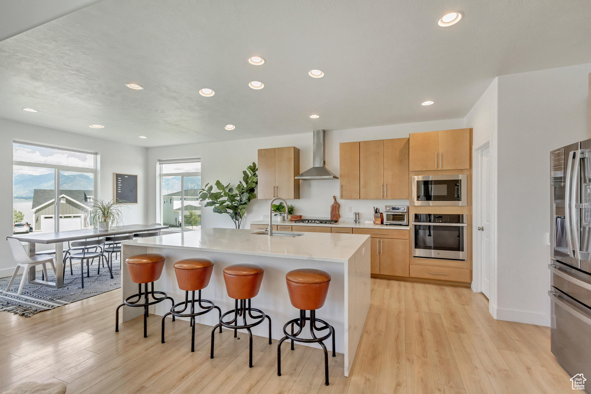 Kitchen with wall chimney range hood, sink, a kitchen island with sink, stainless steel appliances, and light brown cabinets