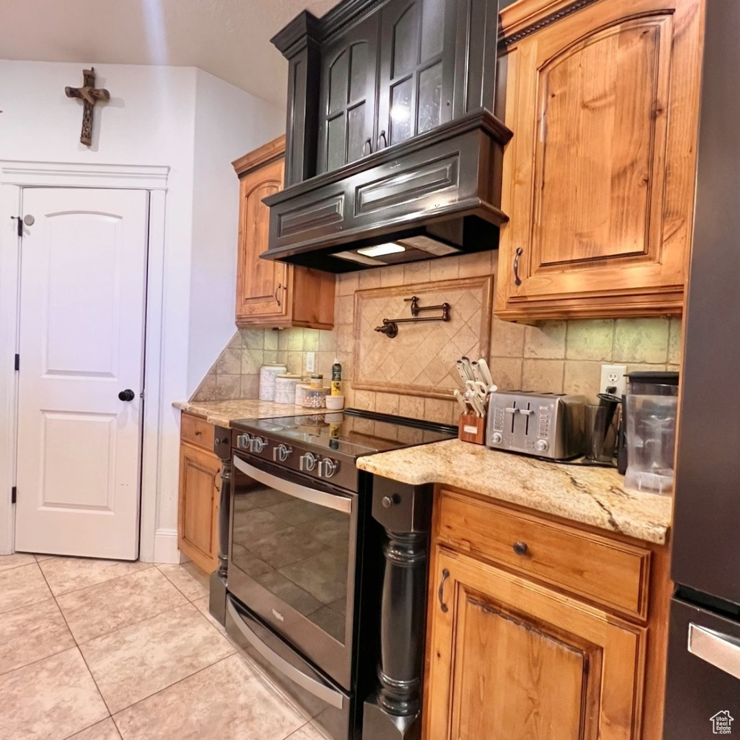 Kitchen featuring light tile patterned flooring, appliances with stainless steel finishes, custom range hood, light stone countertops, and decorative backsplash