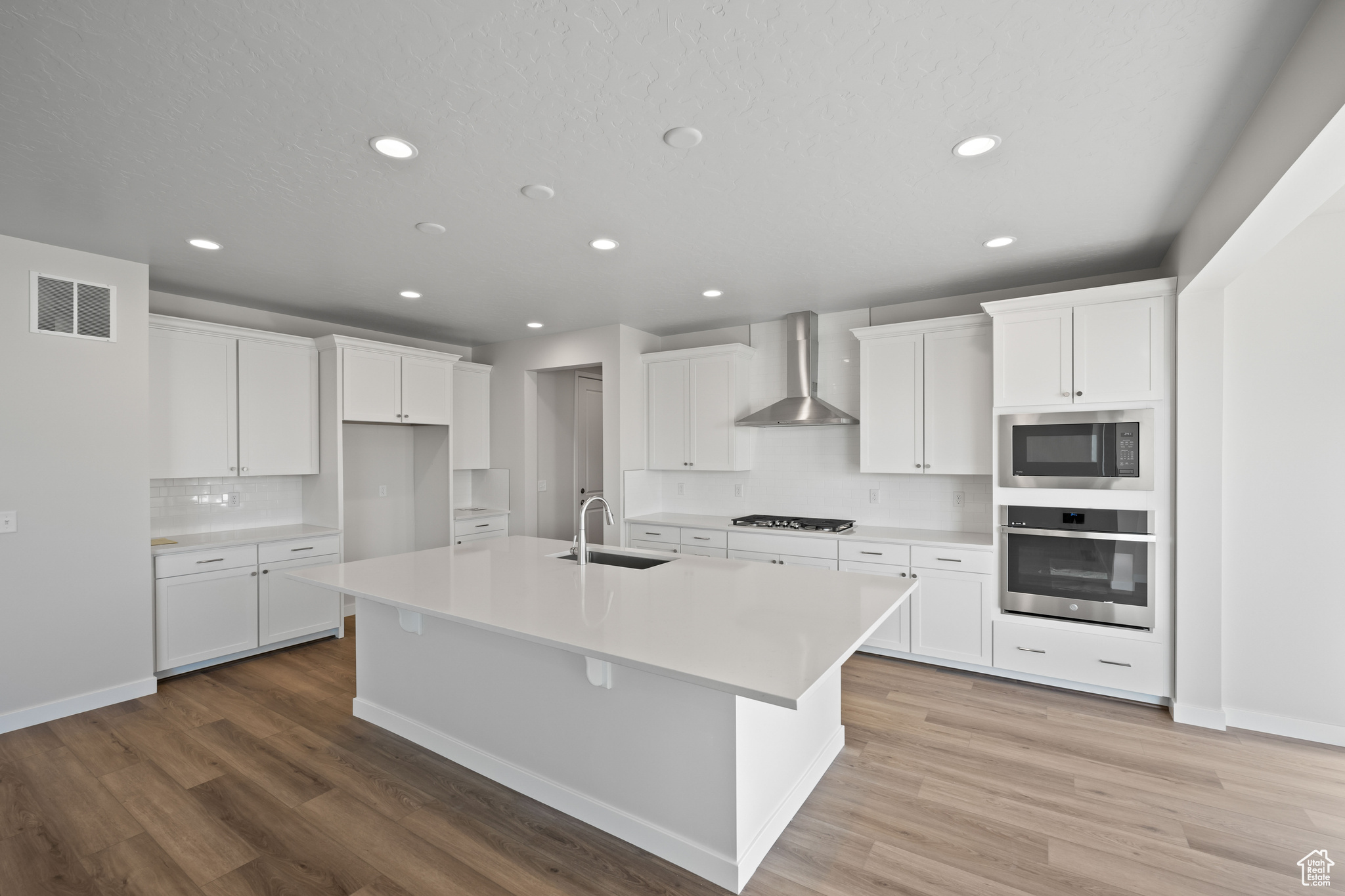 Kitchen with sink, stainless steel oven, an island with sink, wall chimney range hood, and white cabinets